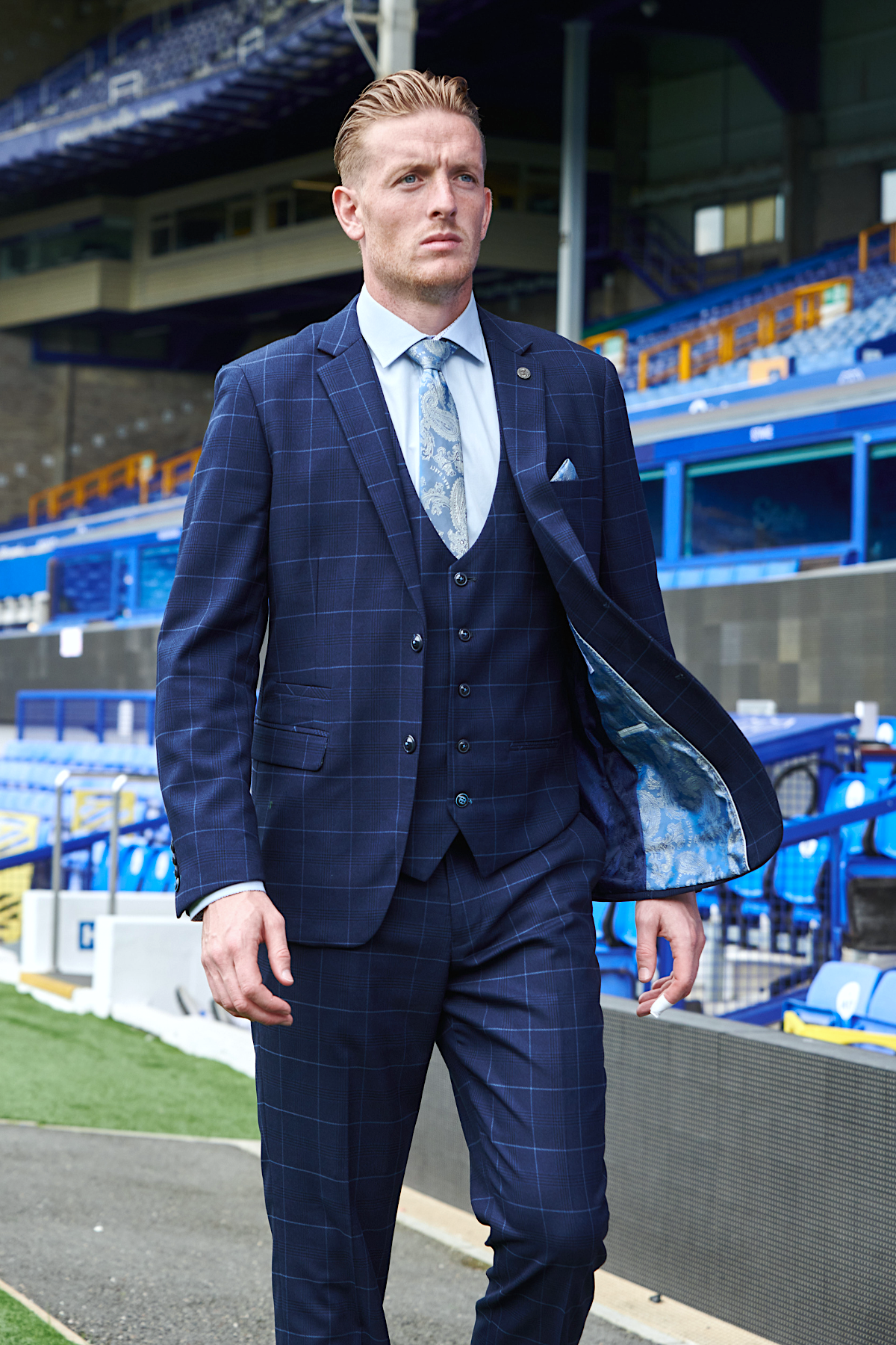 Jordan Pickford wears the Edinson Navy Sky suit from marcdarcy’s Everton Collection, featuring a patterned tie and vest, as he walks confidently in a stadium with blue seats and a large covered stand behind him.