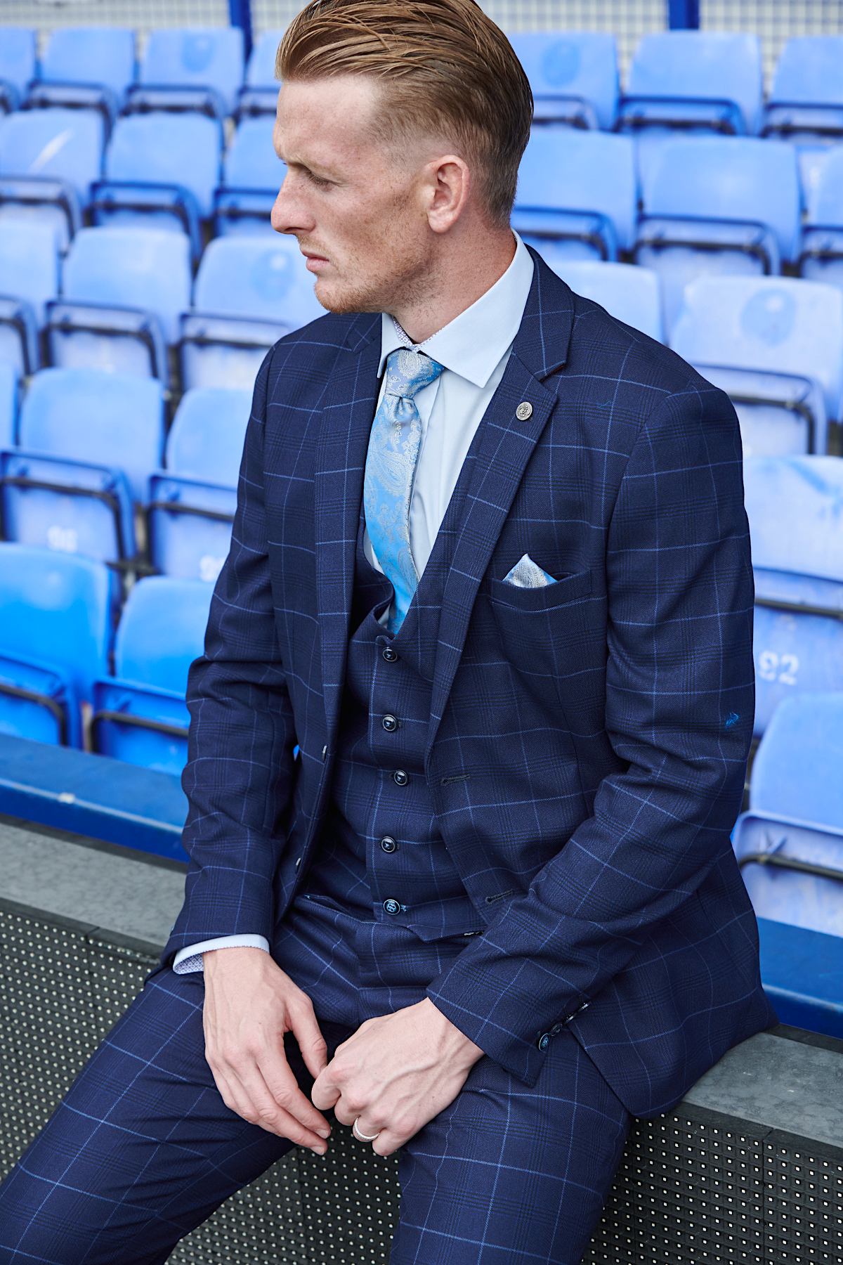 Jordan Pickford, wearing the marcdarcy Edinson Navy Sky suit from The Everton Collection, sits on stadium bleachers.