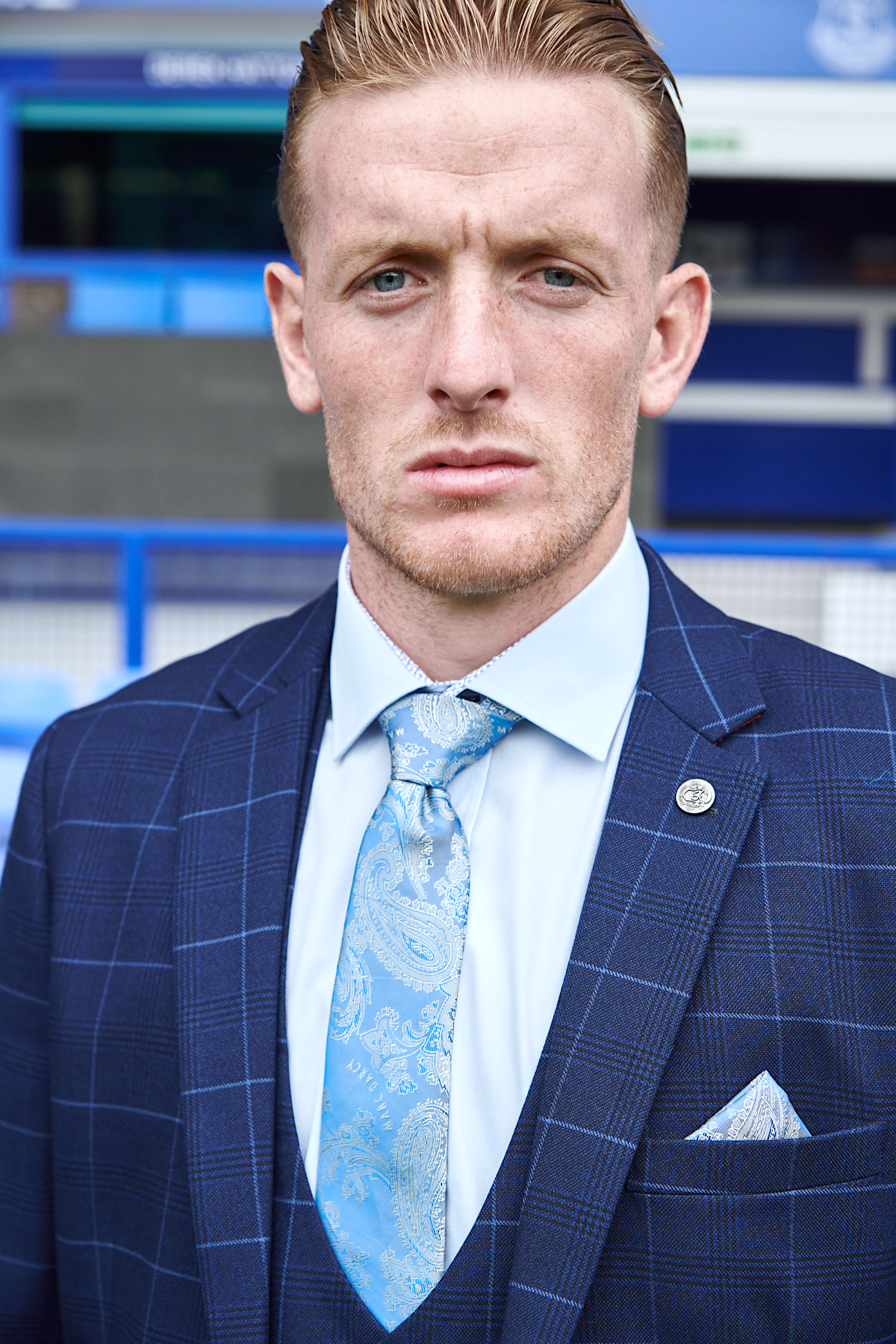 A man wears The Everton Collection | Jordan Pickford in Edinson Navy Sky three-piece suit by marcdarcy, paired with a light blue shirt and patterned blue tie. He stands outdoors with blue stadium seating and barriers behind him, looking serious.