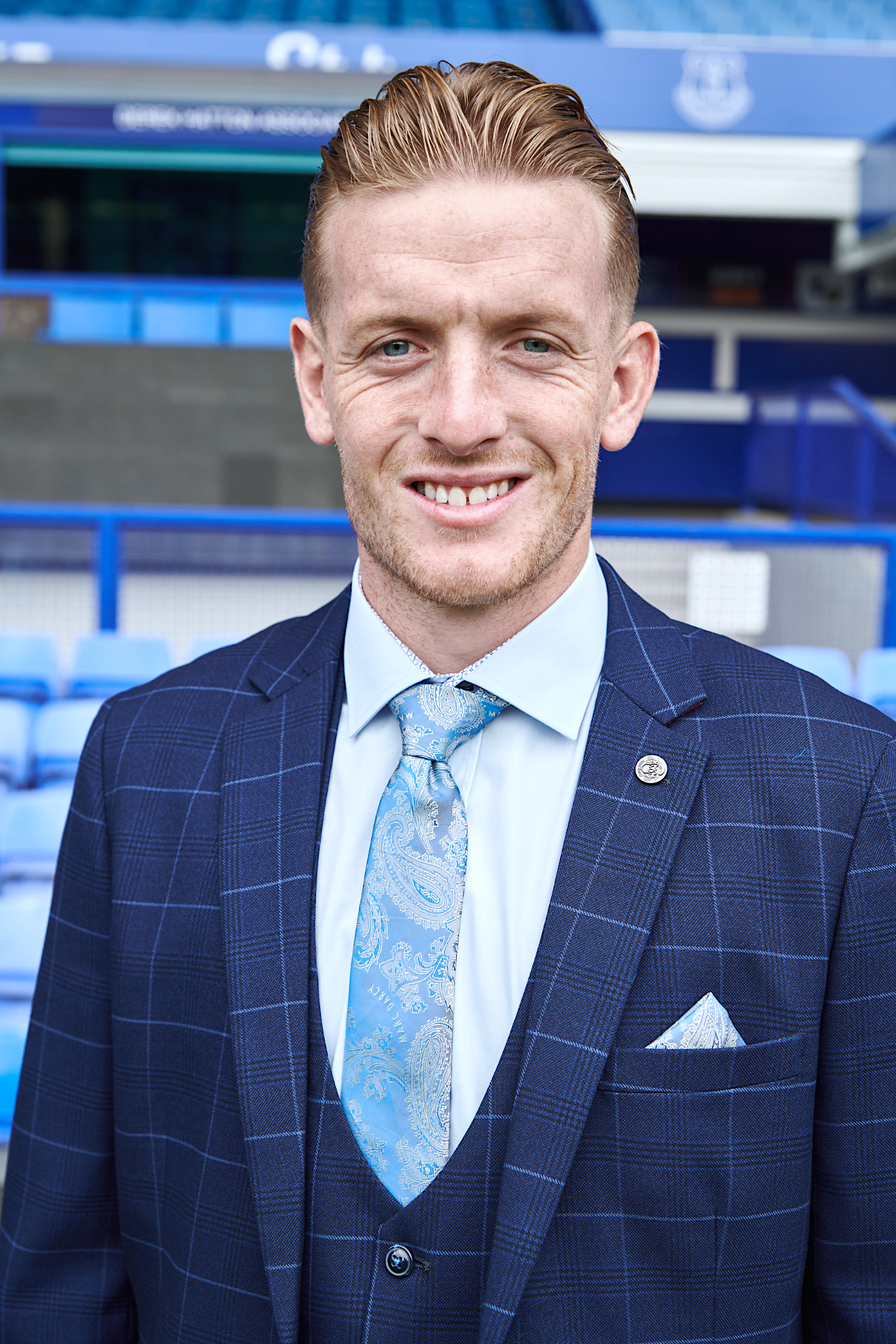 Jordan Pickford smiles at the camera, wearing marcdarcy’s Edinson Navy Sky check suit from The Everton Collection, with a light blue shirt and paisley tie. He stands in front of blue stadium seating.