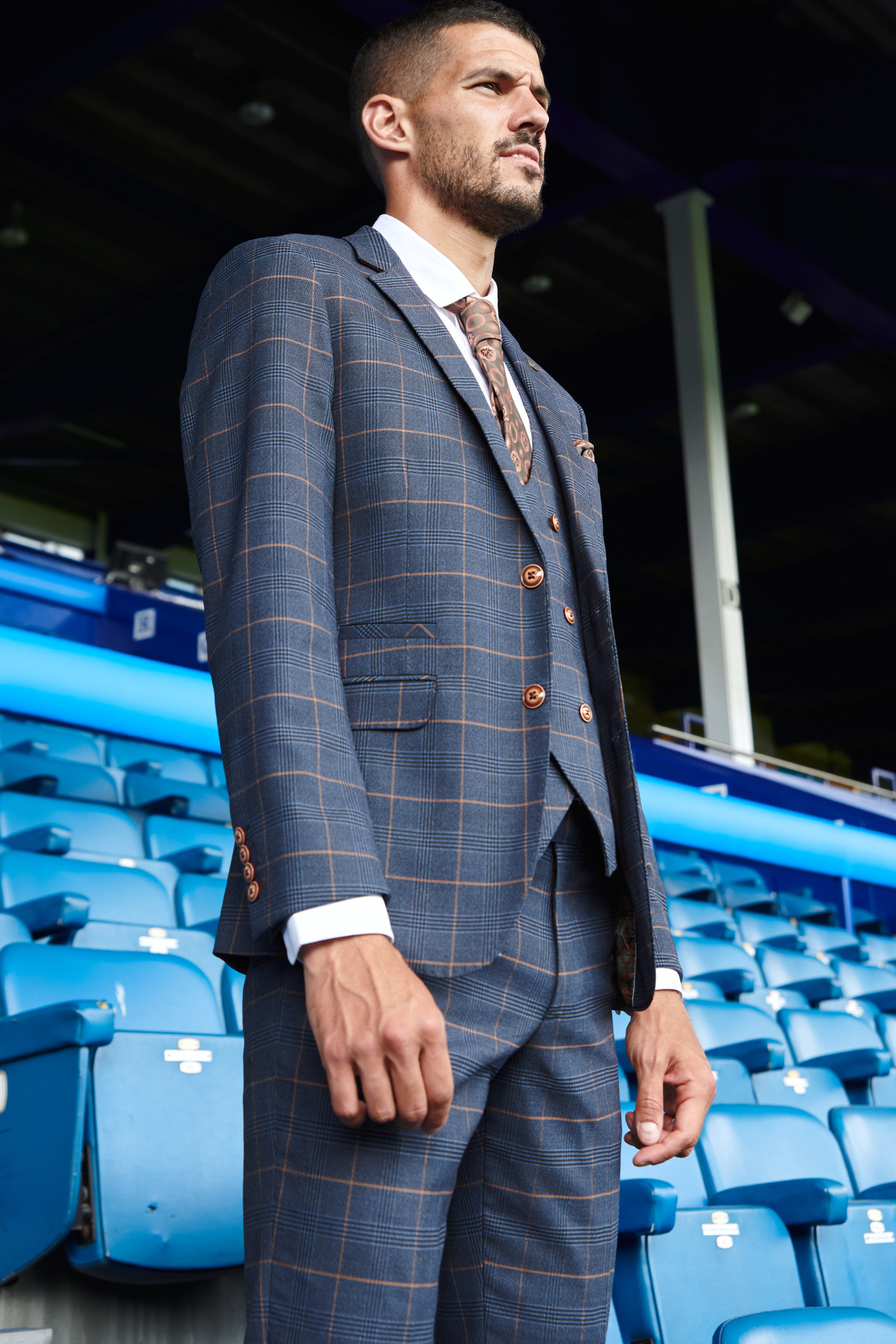 Conor Coady wears the marcdarcy Jenson Marine suit from The Everton Collection, featuring a sharp check pattern and brown tie, standing in a stadium with blue seats and overhead beams behind him.