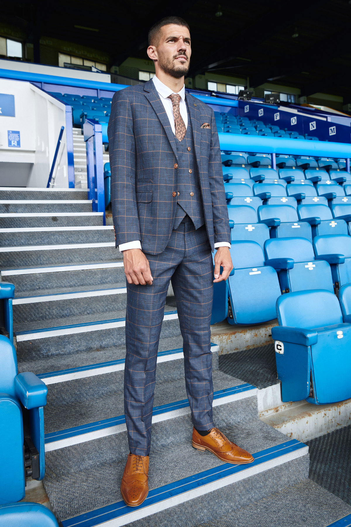 Conor Coady wears marcdarcy’s Jenson Marine slim fit suit with a double breasted waistcoat and brown shoes, standing near blue stadium seats in The Everton Collection.