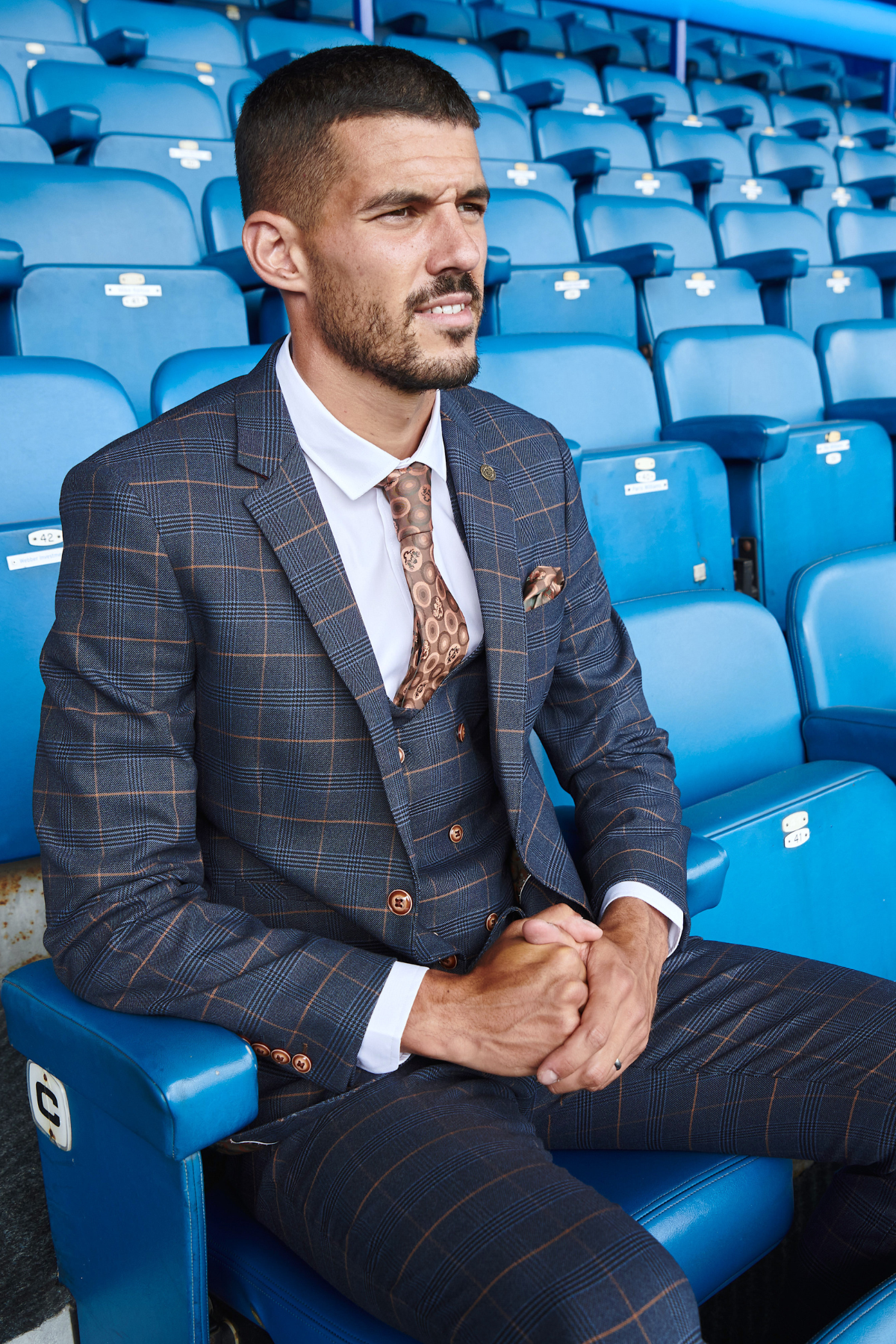 Conor Coady wears the marcdarcy Jenson Marine check suit from The Everton Collection, sitting in a blue stadium seat surrounded by empty seats, hands clasped and looking ahead.