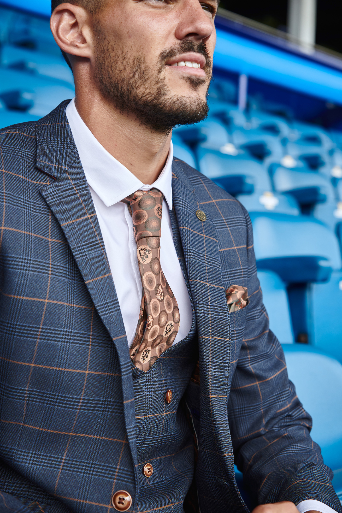 Conor Coady wears marcdarcy's Jenson Marine slim fit blue checkered suit with a brown patterned tie and pocket square from The Everton Collection, sitting in blue stadium seats with a slight smile.