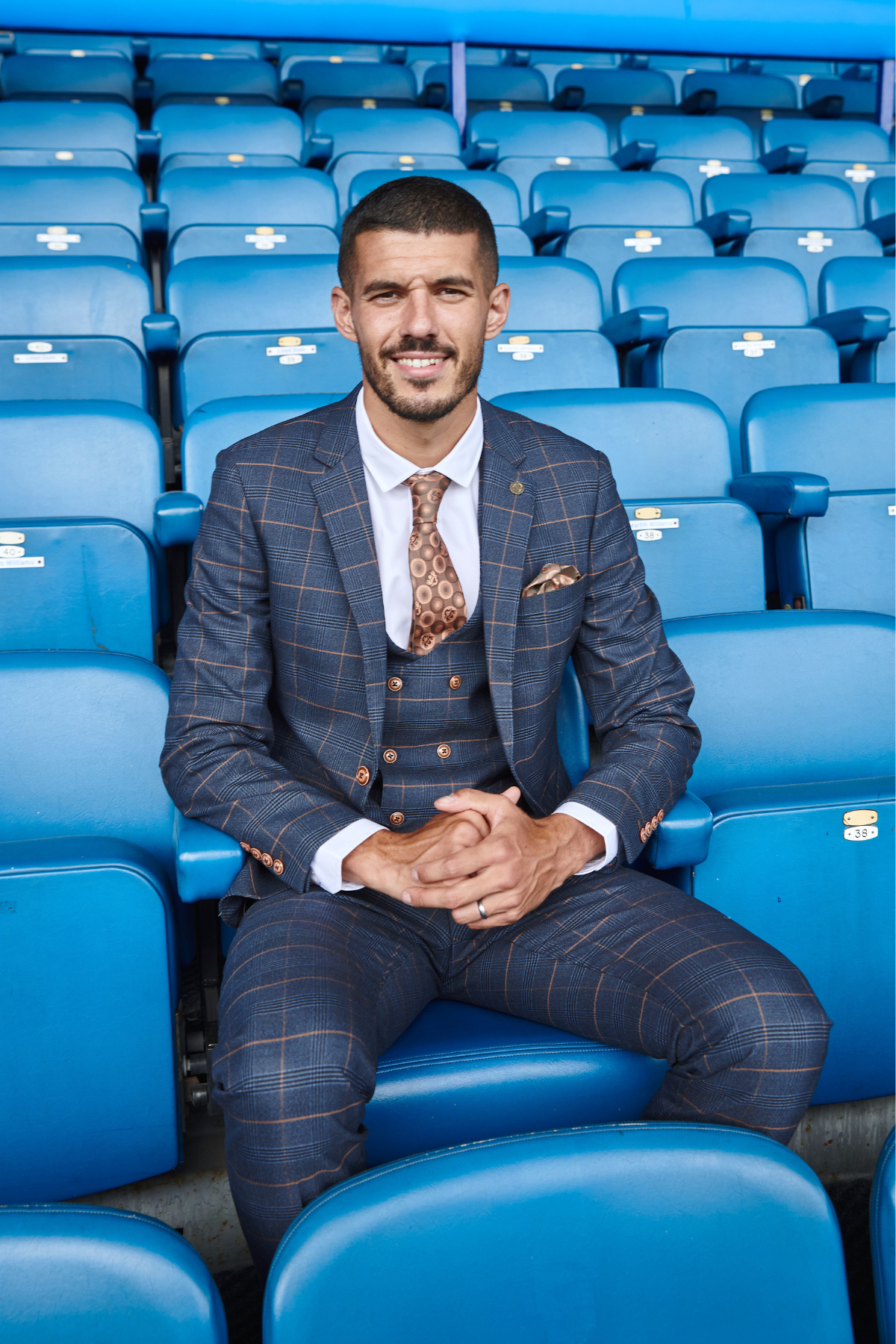 Conor Coady, wearing the marcdarcy Jenson Marine slim fit suit from The Everton Collection, smiles among empty blue Everton F.C. seats—his double breasted waistcoat and brown patterned tie complete the stylish look.