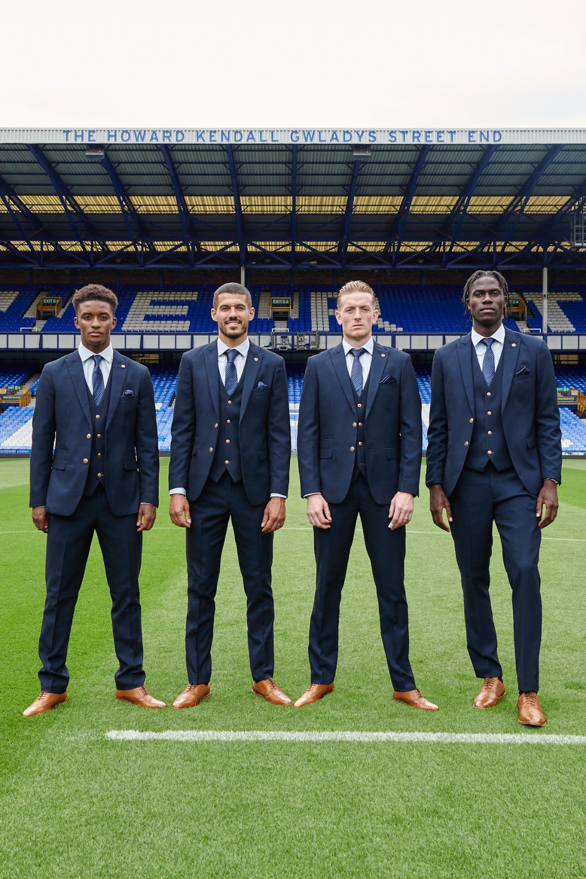 Four men in matching marcdarcy The Everton Collection | Max Navy Team Suits with brown shoes stand side by side on a football field, posing for a photo in front of empty blue stadium seats and THE HOWARD KENDAL GWADYS STREET END sign.