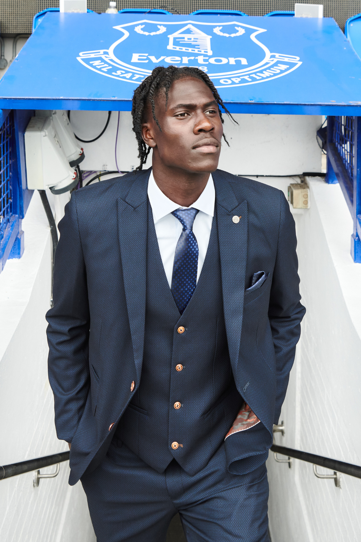A man in The Everton Collection | Max Navy Team Suit by marcdarcy stands at the stadium tunnel entrance beneath the Everton FC crest, wearing a white shirt and blue tie, looking confidently into the distance.