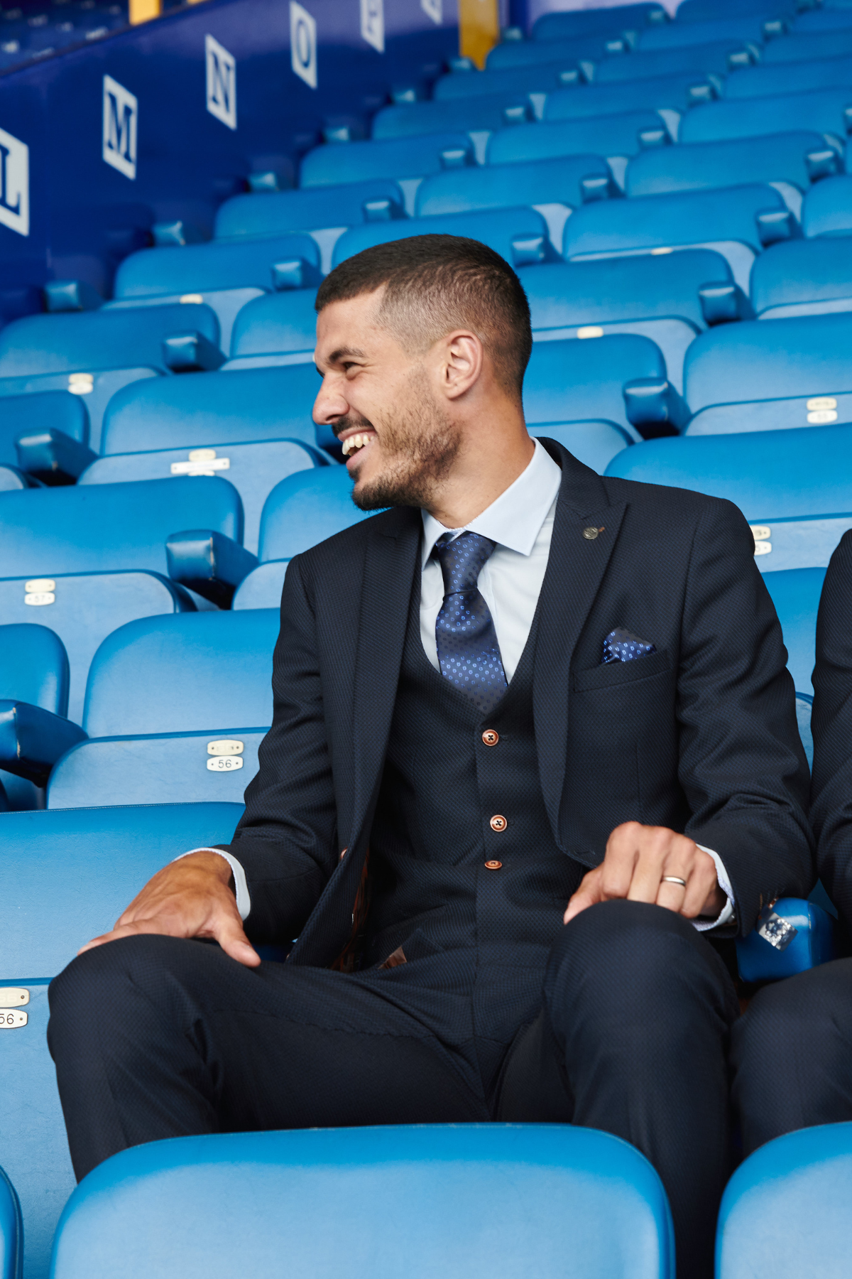 A man in a marcdarcy Max Navy Team Suit from The Everton Collection sits in stadium seats, smiling and looking to his right.
