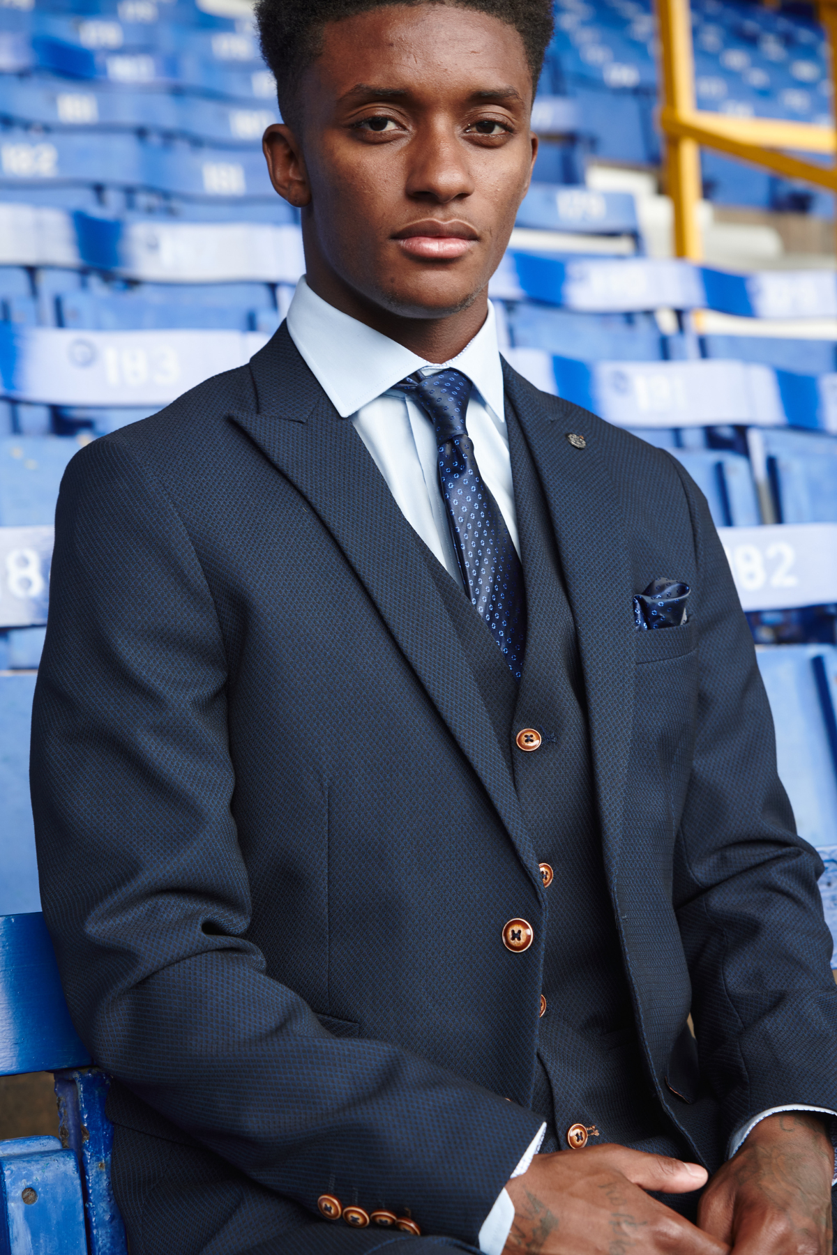A man in a marcdarcy The Everton Collection | Max Navy Team Suit sits confidently on blue stadium seats, wearing a patterned tie and brown buttons. The empty, numbered rows in the background evoke classic formalwear style.