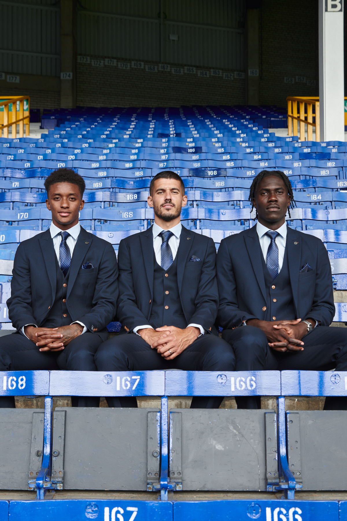 Three men wearing marcdarcy’s The Everton Collection | Max Navy Team Suit sit side by side on blue stadium seats, facing the camera with serious expressions. The empty, numbered seats behind them form a striking pattern.