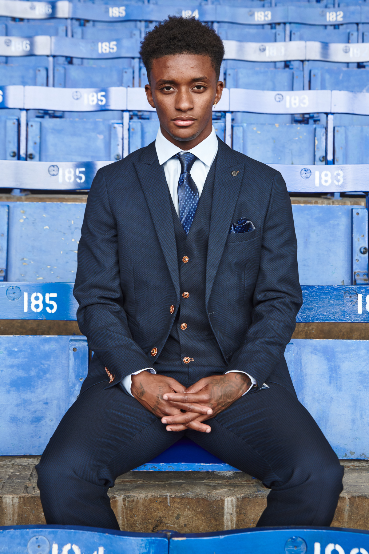 A young man wears the marcdarcy Everton Collection | Max Navy Team Suit as he sits on worn, numbered blue stadium seats, looking seriously at the camera.