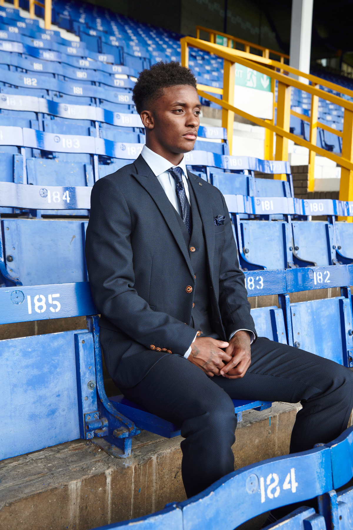 A young Black man in formalwear sits on blue stadium seats, glancing sideways—his marcdarcy Max Navy Team Suit from The Everton Collection striking against the backdrop.