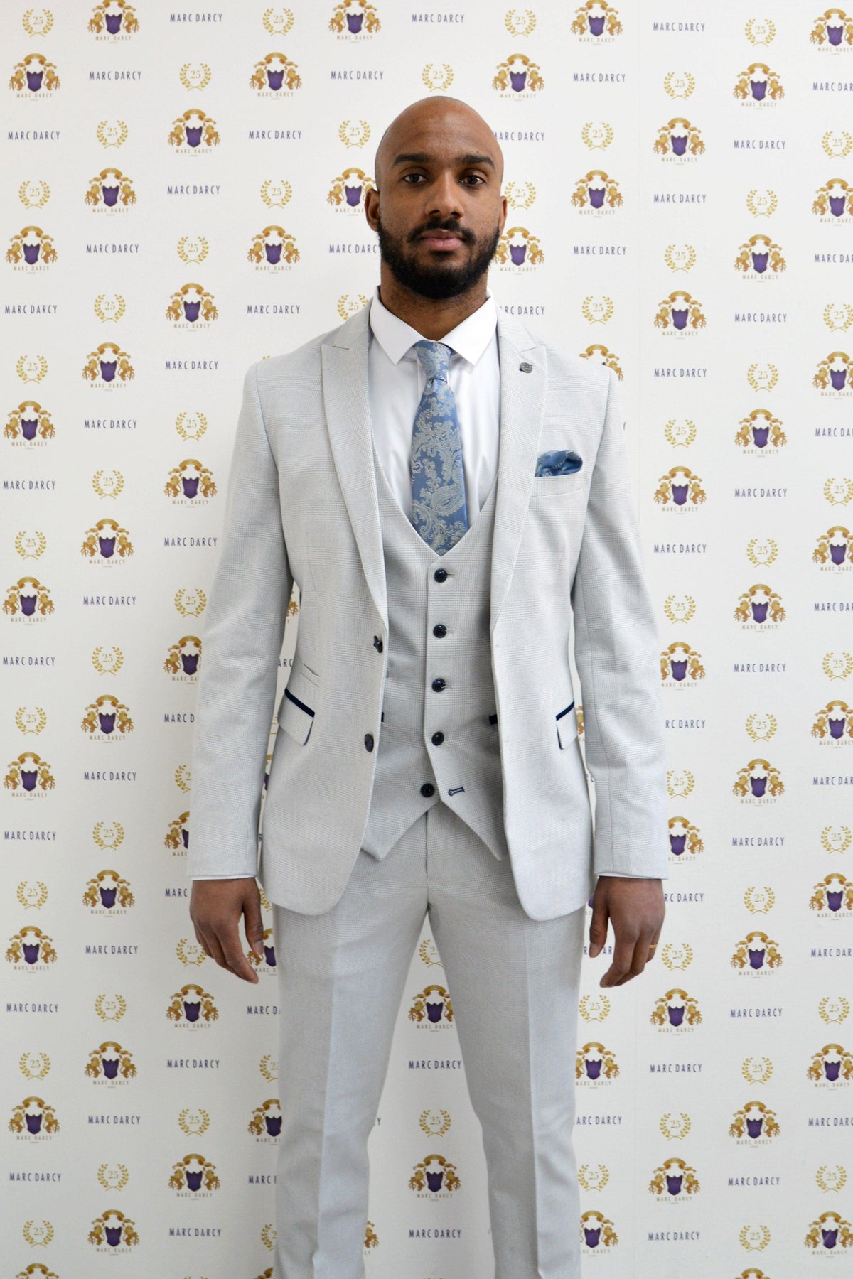 Former Manchester City footballer Fabian Delph wears a BROMLEY stone check suit by Marc Darcy, posing before a backdrop with repeating Marc Darcy logos.