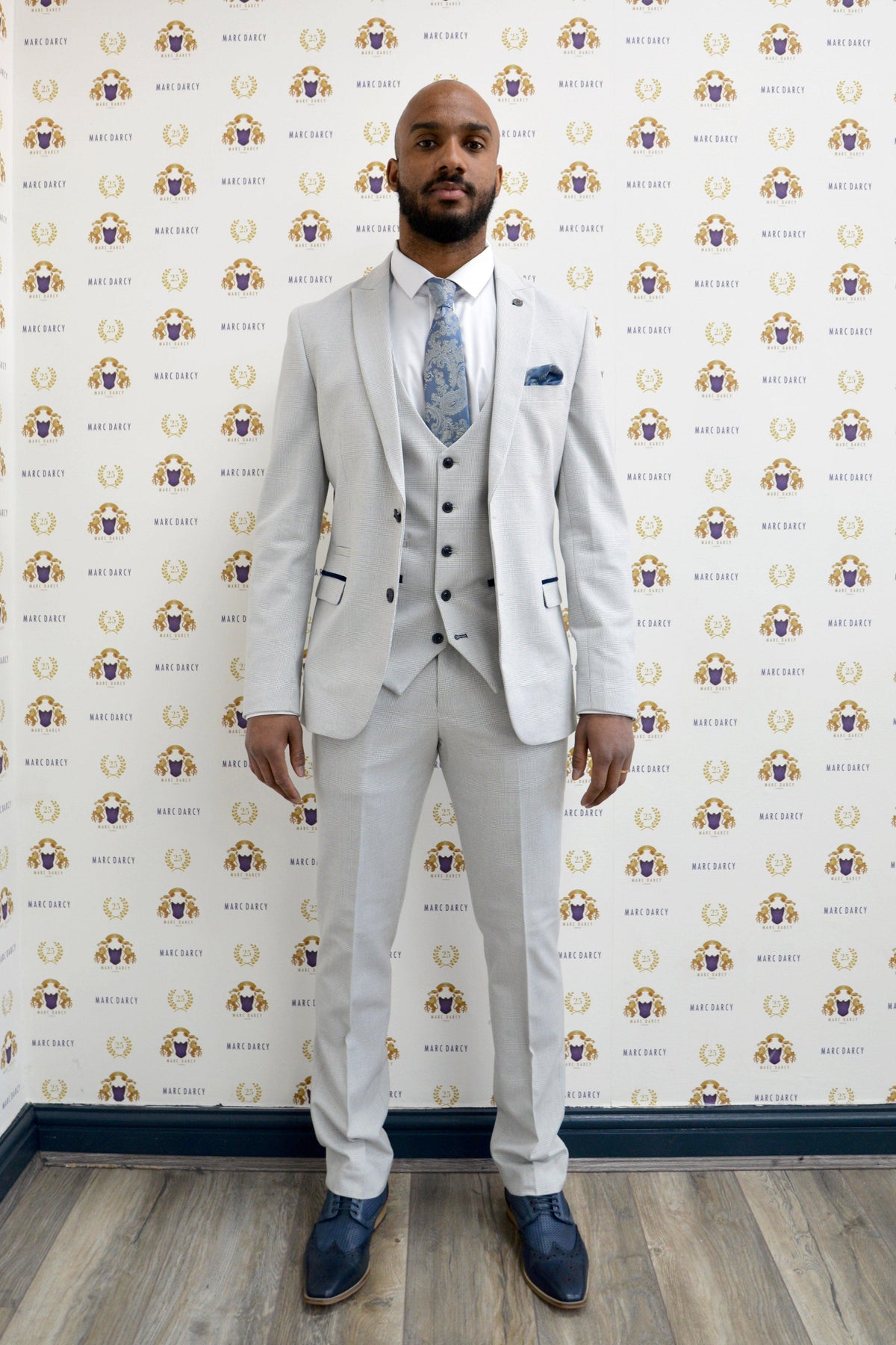 Former Manchester City footballer Fabian Delph wears the Marc Darcy BROMLEY Stone Check Suit, posing confidently against a backdrop with Bare Daisy logos. He pairs it with a white shirt, patterned tie, dark shoes, and sports short hair with a beard.