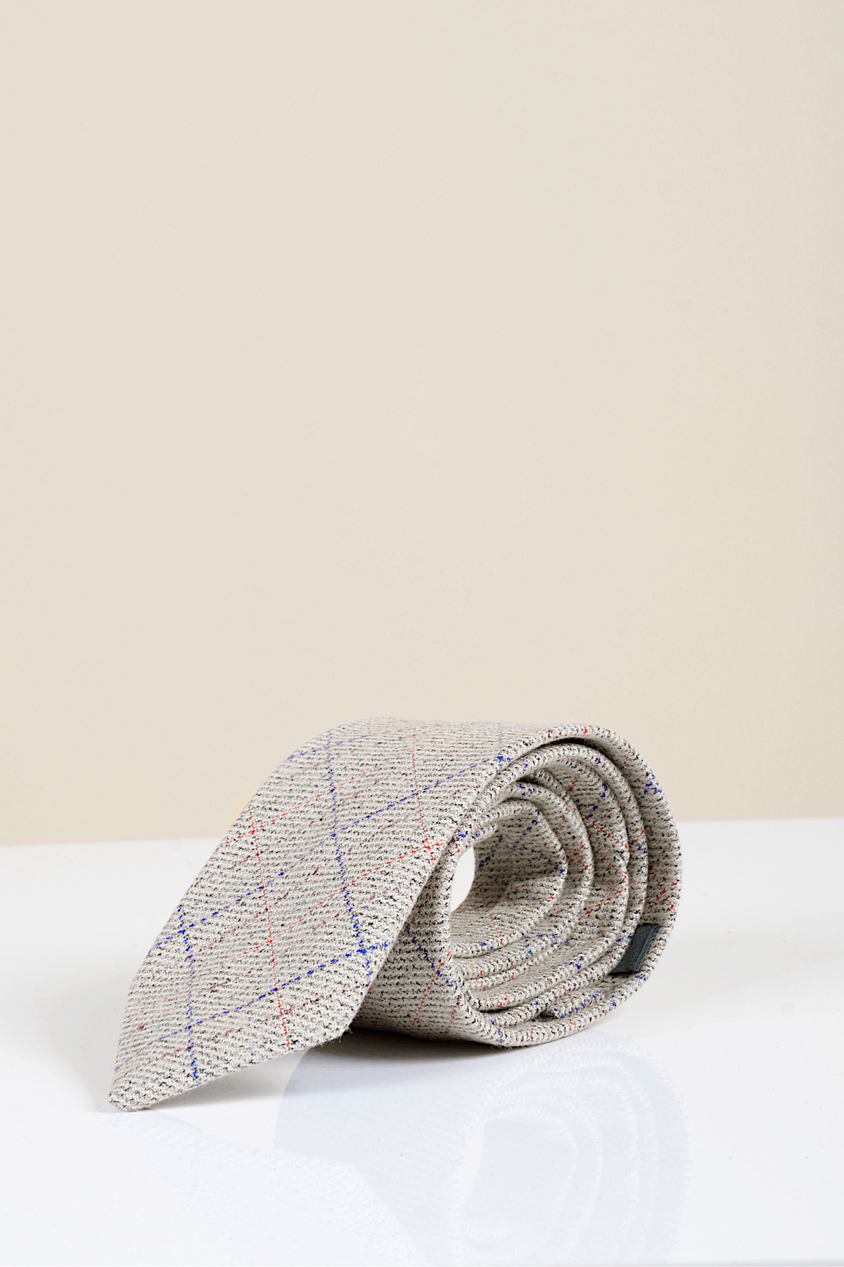 The Harding - Cream Tweed Check Tie by Accessory features a cream base with blue and red checks, showcased rolled up on a glossy white surface against a plain beige background.