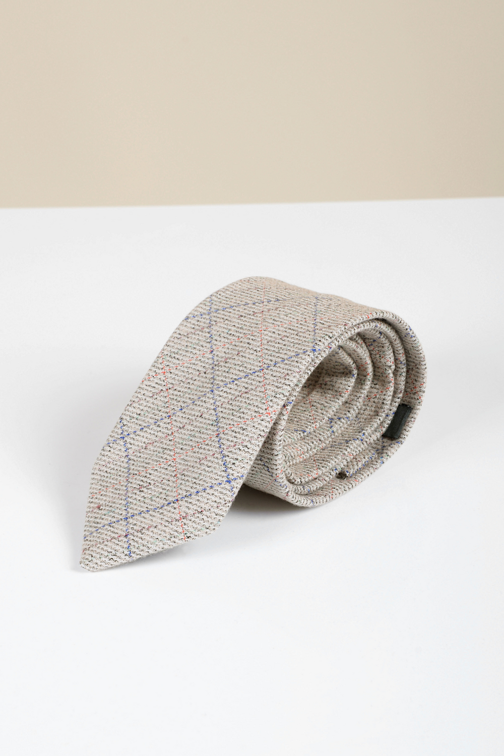 The Accessory Harding - Cream Tweed Check Tie, featuring a beige herringbone pattern with blue and red plaid lines, is rolled up on a white surface against a light beige background.