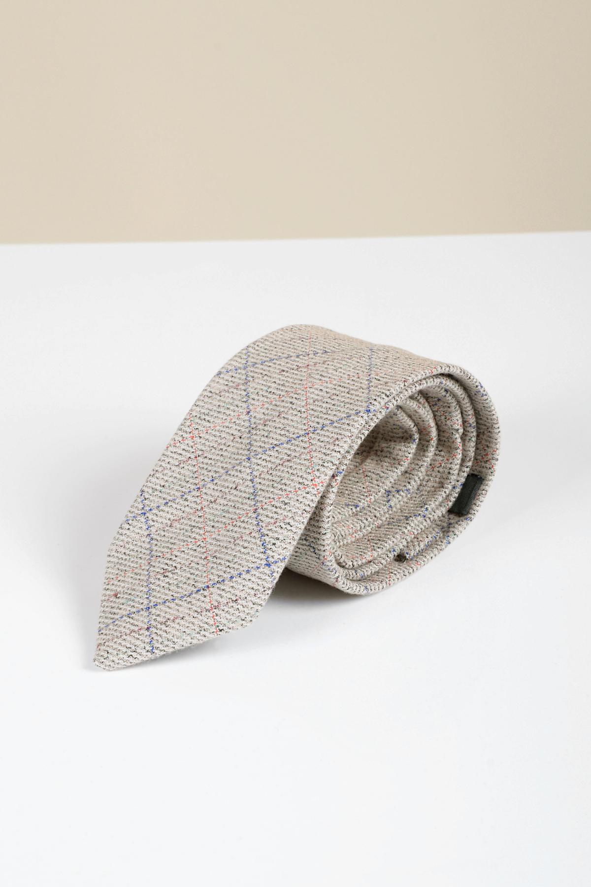 The Accessory Harding - Cream Tweed Check Tie, featuring a beige herringbone pattern with blue and red plaid lines, is rolled up on a white surface against a light beige background.