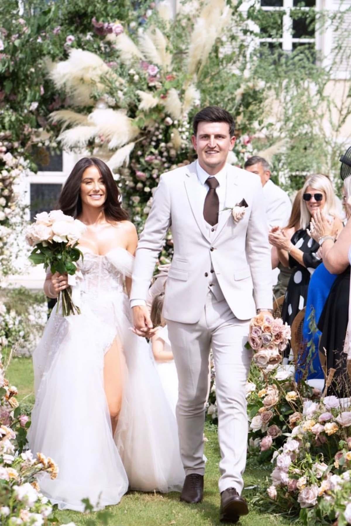 A bride in a strapless white gown and a groom wearing the Marc Darcy HM5 - Stone Tailored Three Piece Suit walk hand in hand outdoors among flowers, as guests smile and applaud.