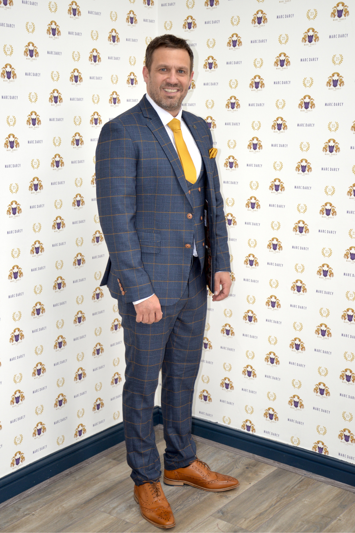 Actor Jamie Lomas smiles in front of a wall with repeating “marcdarcy” logos, wearing the Jenson Marine Check Suit by marcdarcy, paired with a yellow tie and brown shoes.