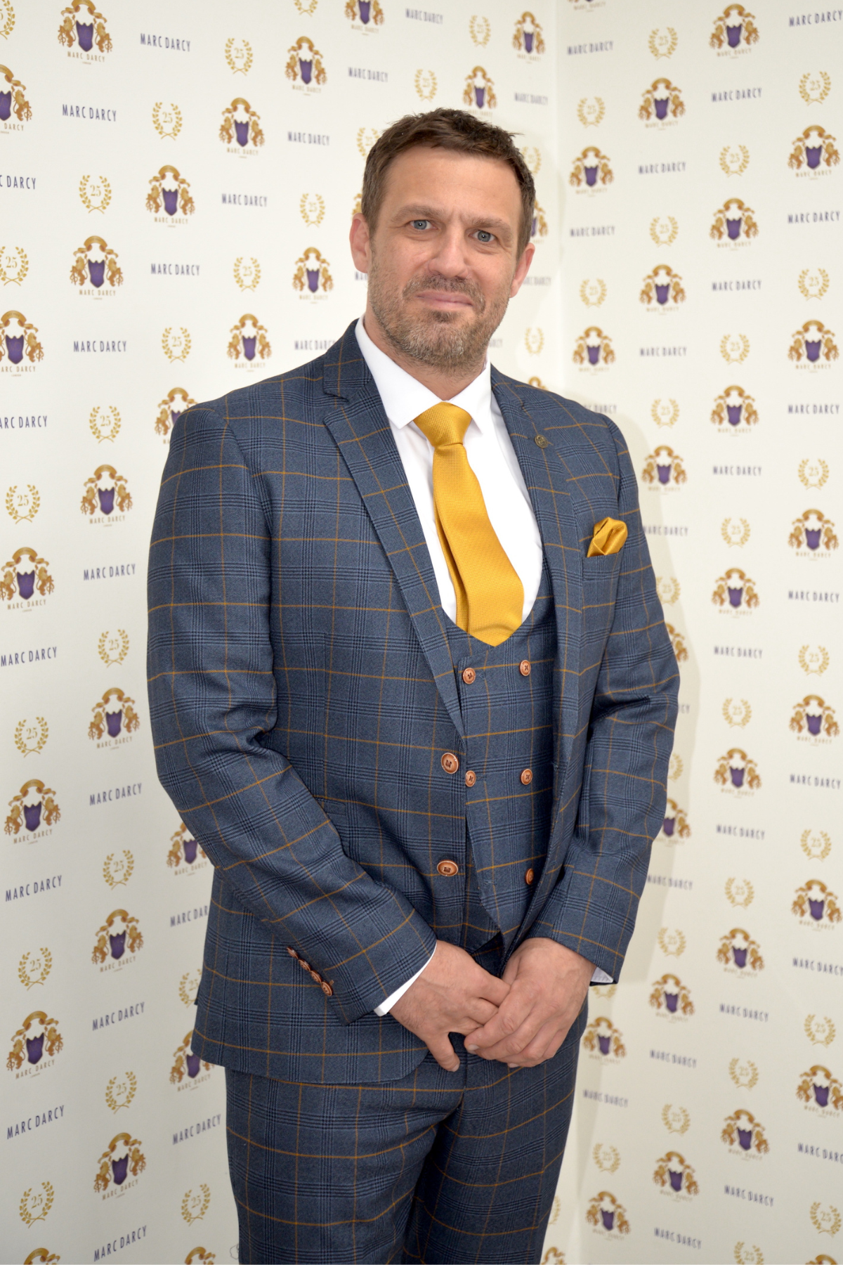 Actor Jamie Lomas stands before a marcdarcy backdrop wearing the Jenson Marine Check Suit, styled with a yellow tie and pocket square, hands clasped and smiling slightly.