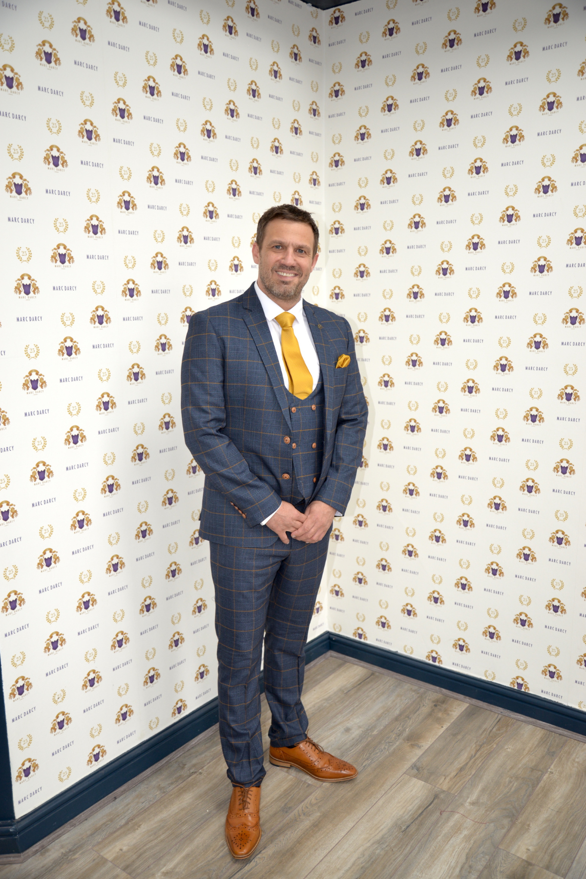 Actor Jamie Lomas wears the marcdarcy Jenson Marine Check Suit with a yellow tie and brown shoes, smiling in front of a wall featuring repeating marcdarcy logos.