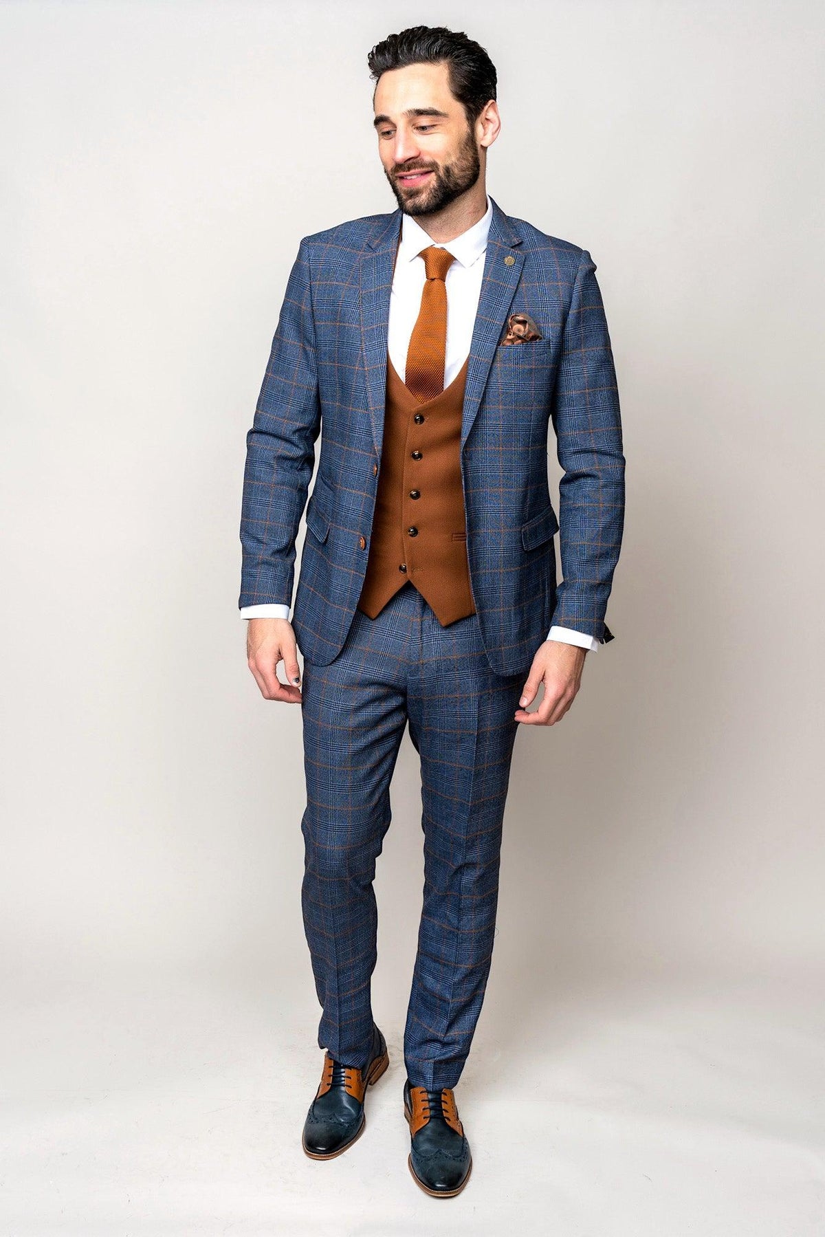 A man with dark hair and a trimmed beard stands against a plain background wearing the marcdarcy JENSON Sky Blue Check Suit with Kelvin Tan Waistcoat, styled with a brown tie, pocket square, and coordinating blue and brown dress shoes.