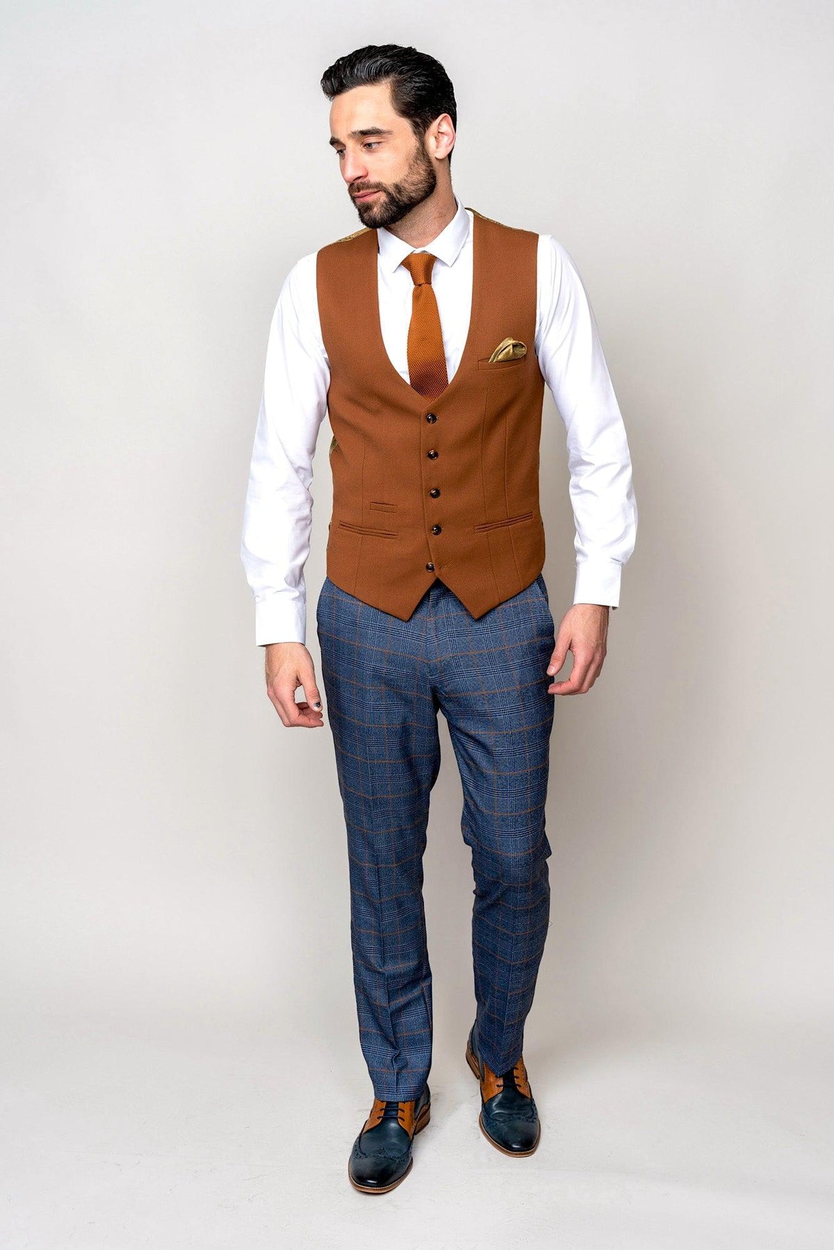 A man wears the marcdarcy JENSON Sky Blue Check Suit paired with the Kelvin Tan Waistcoat, styled with a white shirt and brown tie. He stands against a light background, looking left, one hand by his side and the other near his pocket.