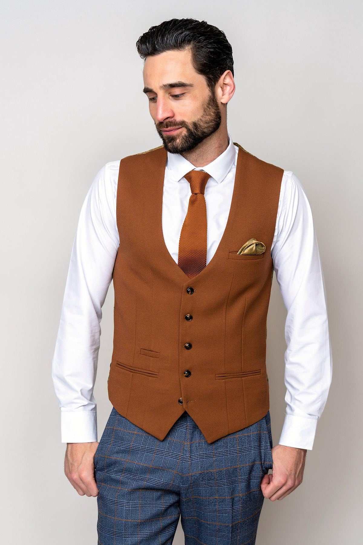 A man with dark hair and a beard wears the marcdarcy JENSON - Sky Blue Check Suit paired with a Kelvin Tan Waistcoat, white shirt, tie, and yellow pocket square. He looks downward with his hands by his sides.