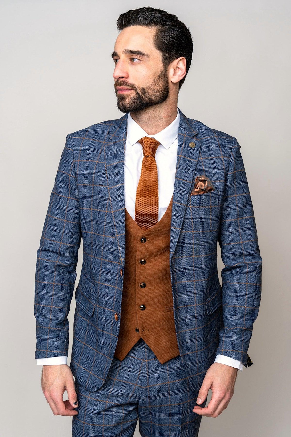 A dark-haired, bearded man wears the marcdarcy JENSON Sky Blue Check Suit with the Kelvin Tan Waistcoat, paired with a tie and pocket square, standing against a plain light background while looking to his left.