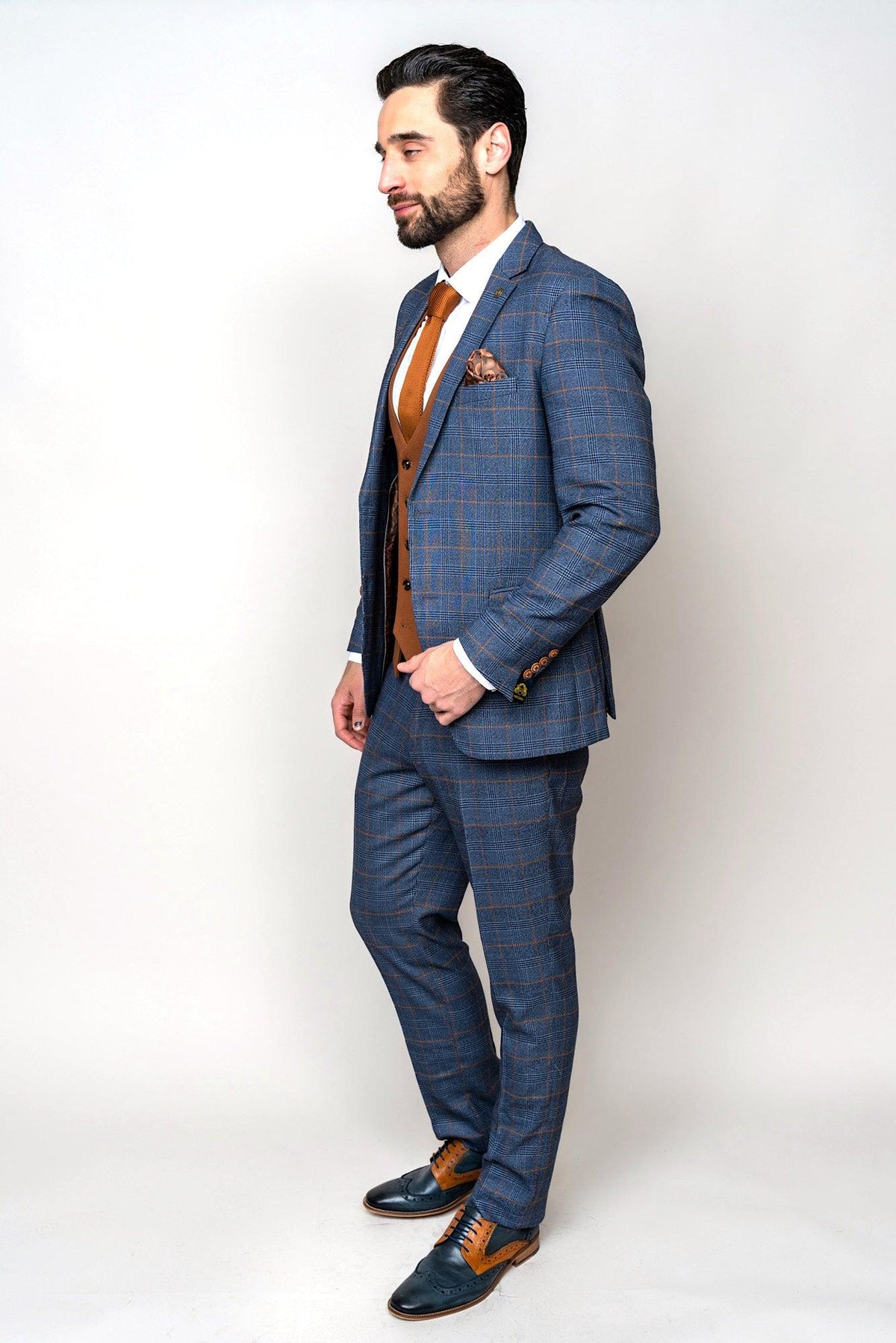 A man stands in profile wearing the marcdarcy JENSON Sky Blue Check Suit with a Kelvin Tan Waistcoat, white shirt, patterned pocket square, and two-tone brown and blue dress shoes against a plain light background.