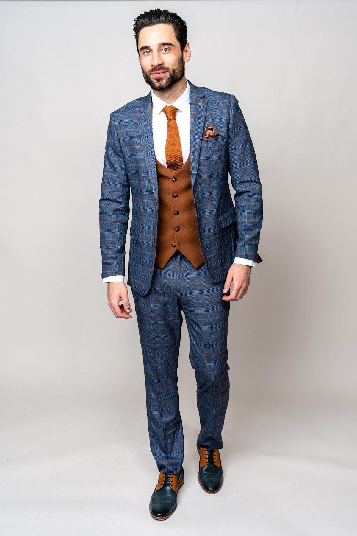 A man in a marcdarcy JENSON Sky Blue Check Suit with a Kelvin Tan Waistcoat and orange tie stands against a light gray background.