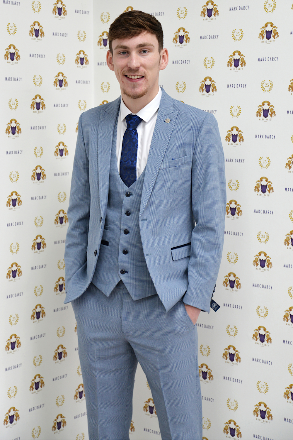 Everton midfielder James Garner stands smiling in front of a gold and purple marcdarcy backdrop, wearing the Bromley Sky Three Piece Suit for a stylish look completed with a coordinating blue tie.