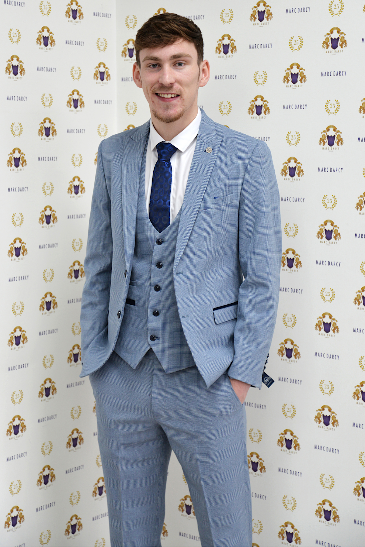 Everton midfielder James Garner stands smiling in front of a gold and purple marcdarcy backdrop, wearing the Bromley Sky Three Piece Suit for a stylish look completed with a coordinating blue tie.