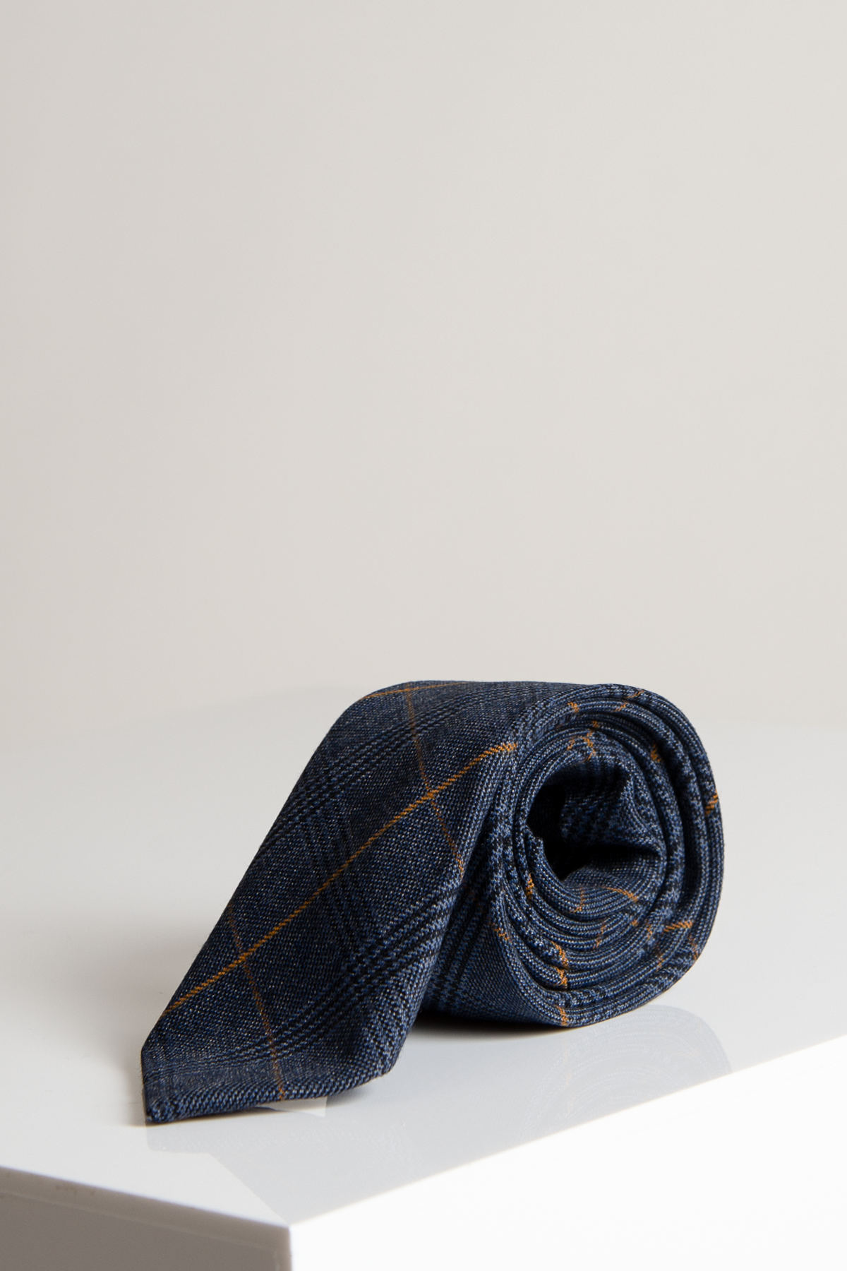 The JENSON - Marine Navy Check Tie by Marc Darcy, featuring thin orange lines, is displayed on a glossy white surface and pairs perfectly with a Kelvin Tan Waistcoat against a plain light background.