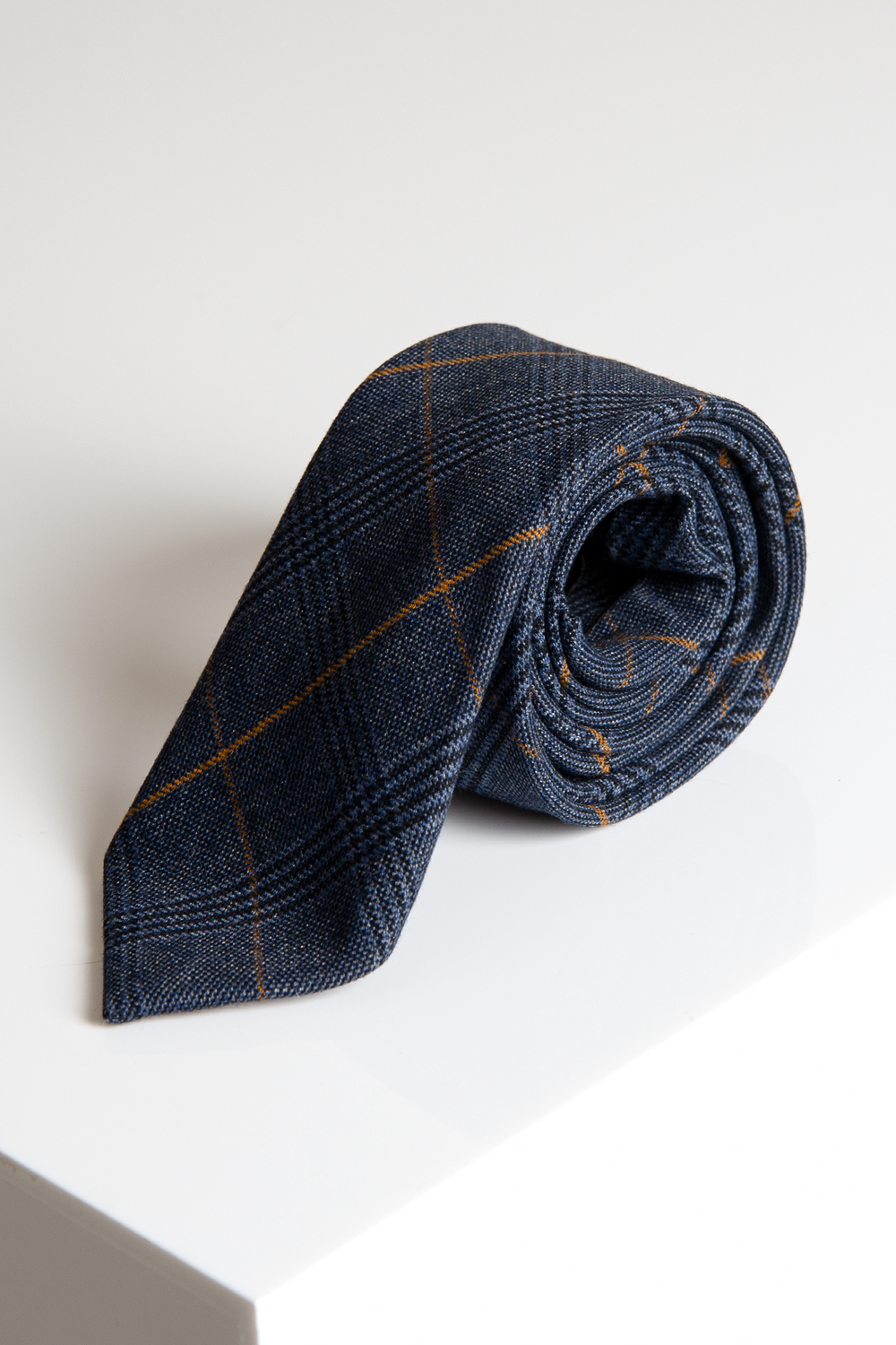 A rolled-up Marc Darcy JENSON - Marine Navy Check Tie featuring thin orange lines is displayed on a white surface.