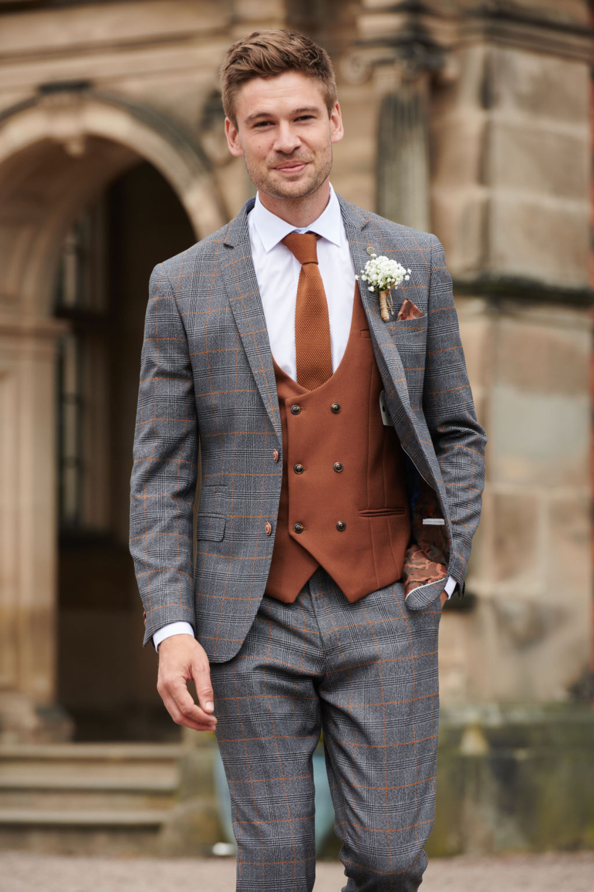 A man dressed in a marcdarcy JENSON Grey Check Suit with a KELVIN Tan Waistcoat, white shirt, brown tie, and boutonniere stands smiling outside a historic stone building.