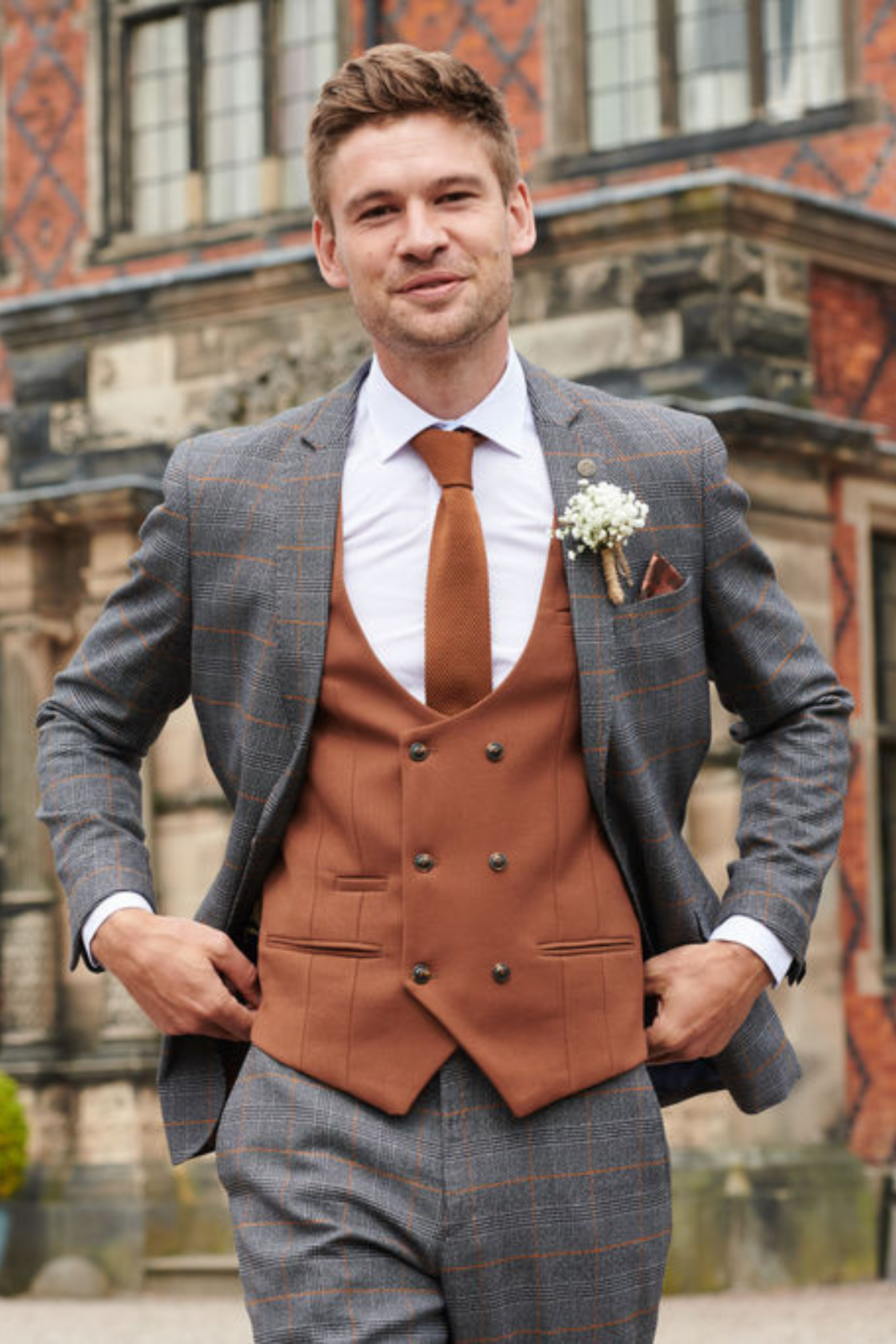 A man, smiling outdoors with a boutonnière on his lapel, wears the marcdarcy JENSON grey check suit and KELVIN tan waistcoat; a historic building stands in the background.