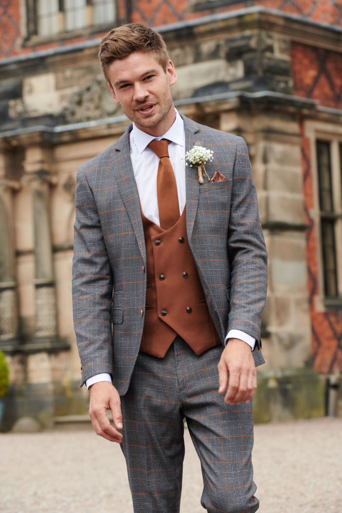 A groom smiles in front of a stone building, wearing the marcdarcy JENSON Grey Check Suit with a KELVIN Tan Waistcoat, matching tie, boutonnière, and pocket square.