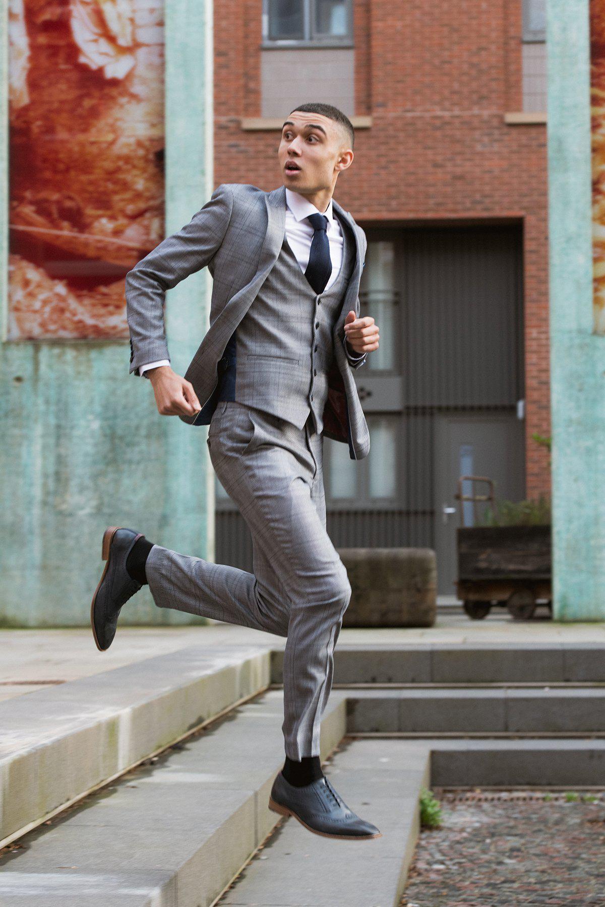 A young man wearing the marcdarcy JERRY Grey Check Suit with Single Breasted Waistcoat, white shirt, and black tie runs up outdoor concrete steps, glancing back in surprise, with brick and concrete buildings behind him.