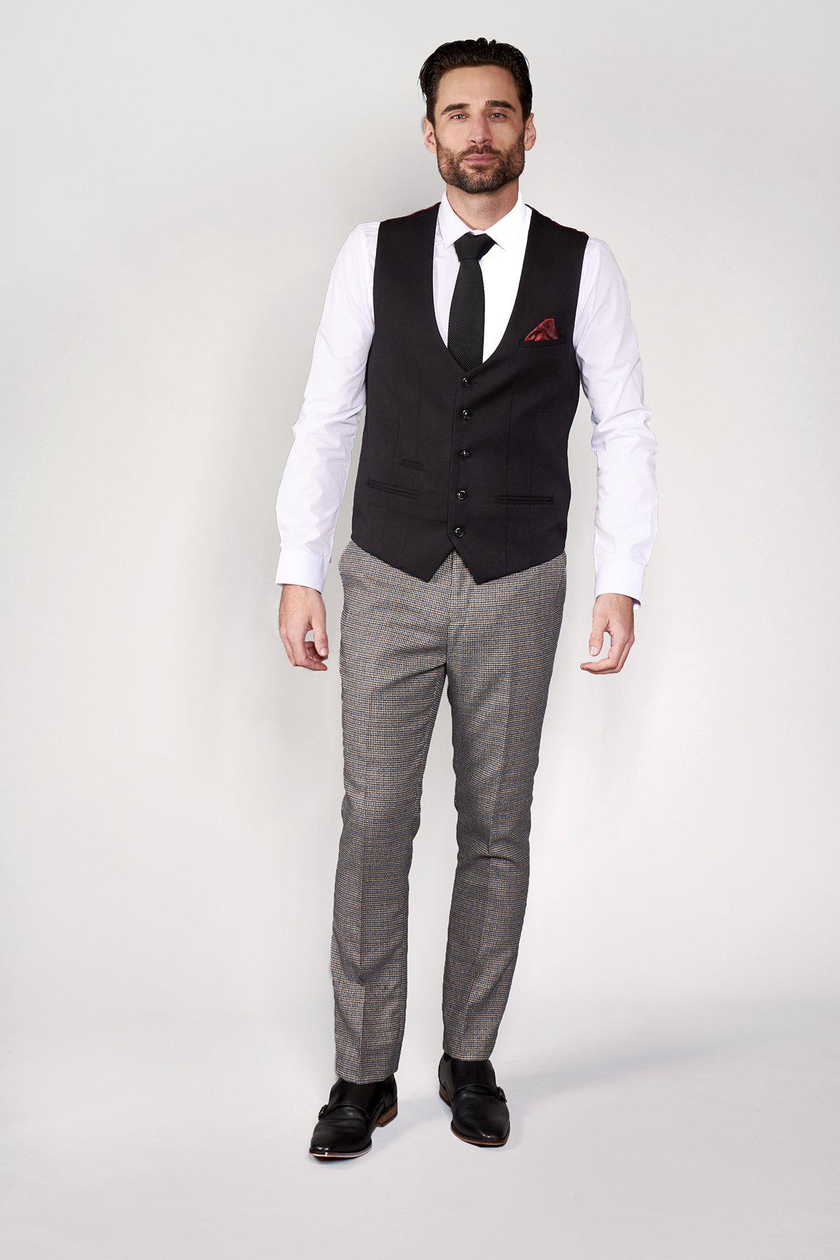 A man wears formal attire against a light background: marcdarcys KELLY Black Single Breasted Waistcoat with a red pocket square, white dress shirt, black tie, gray patterned trousers, and black shoes.