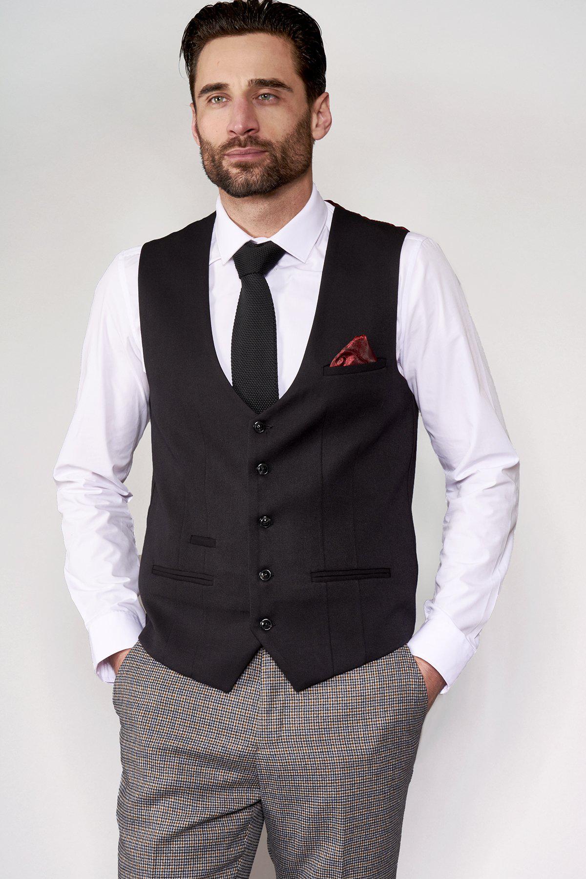 A man models the marcdarcy KELLY black single breasted waistcoat with a white shirt, black tie, and patterned trousers, hands in pockets, highlighting a modern formalwear look.