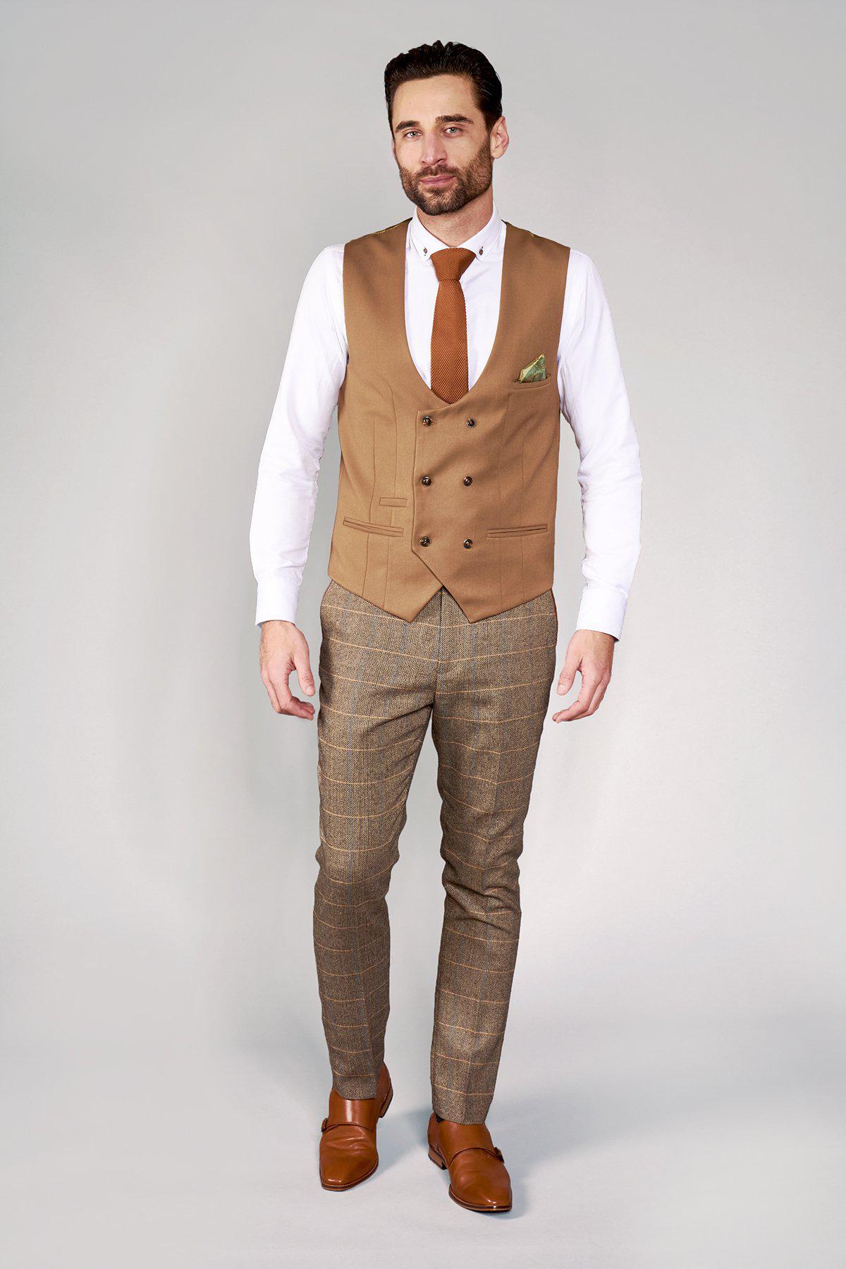 A man in the marcdarcy KELLY Tan Double Breasted Waistcoat, white shirt, brown tie, checked trousers, and brown shoes stands against a light background. He has short dark hair, a beard, and gazes confidently at the camera.