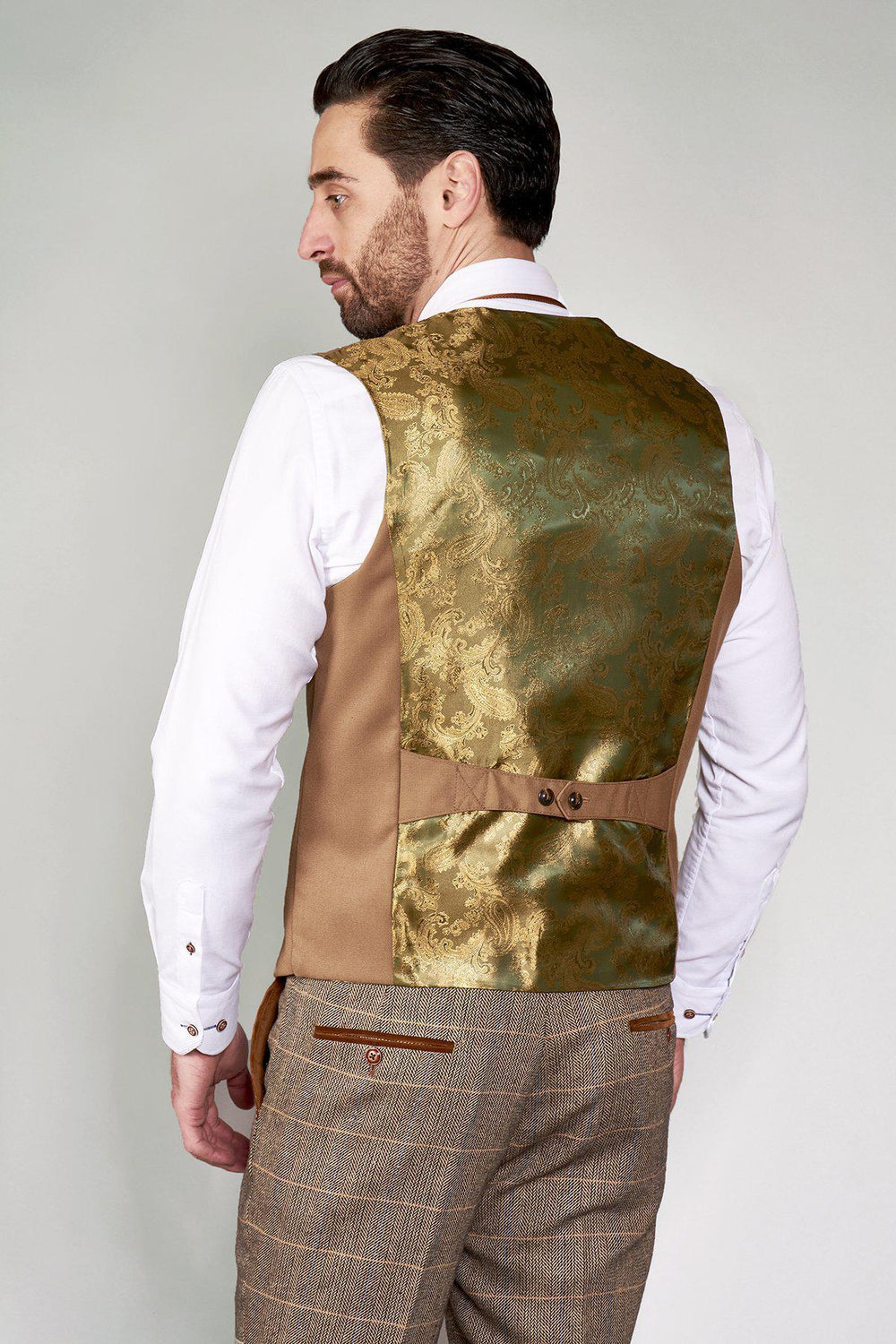 A man with dark hair and beard wears a white shirt, brown patterned trousers, and the KELLY Tan Double Breasted Waistcoat by marcdarcy, featuring gold brocade and an adjustable back strap, showcasing refined British tailoring.