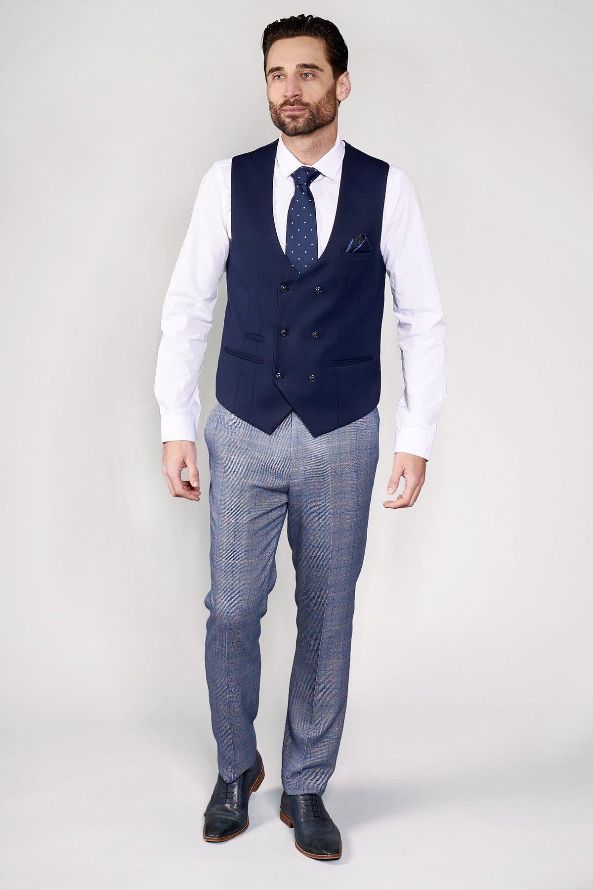 A man wearing the marcdarcy KELLY blue double breasted waistcoat, white shirt, blue patterned tie, blue plaid trousers, and black shoes stands against a light background.