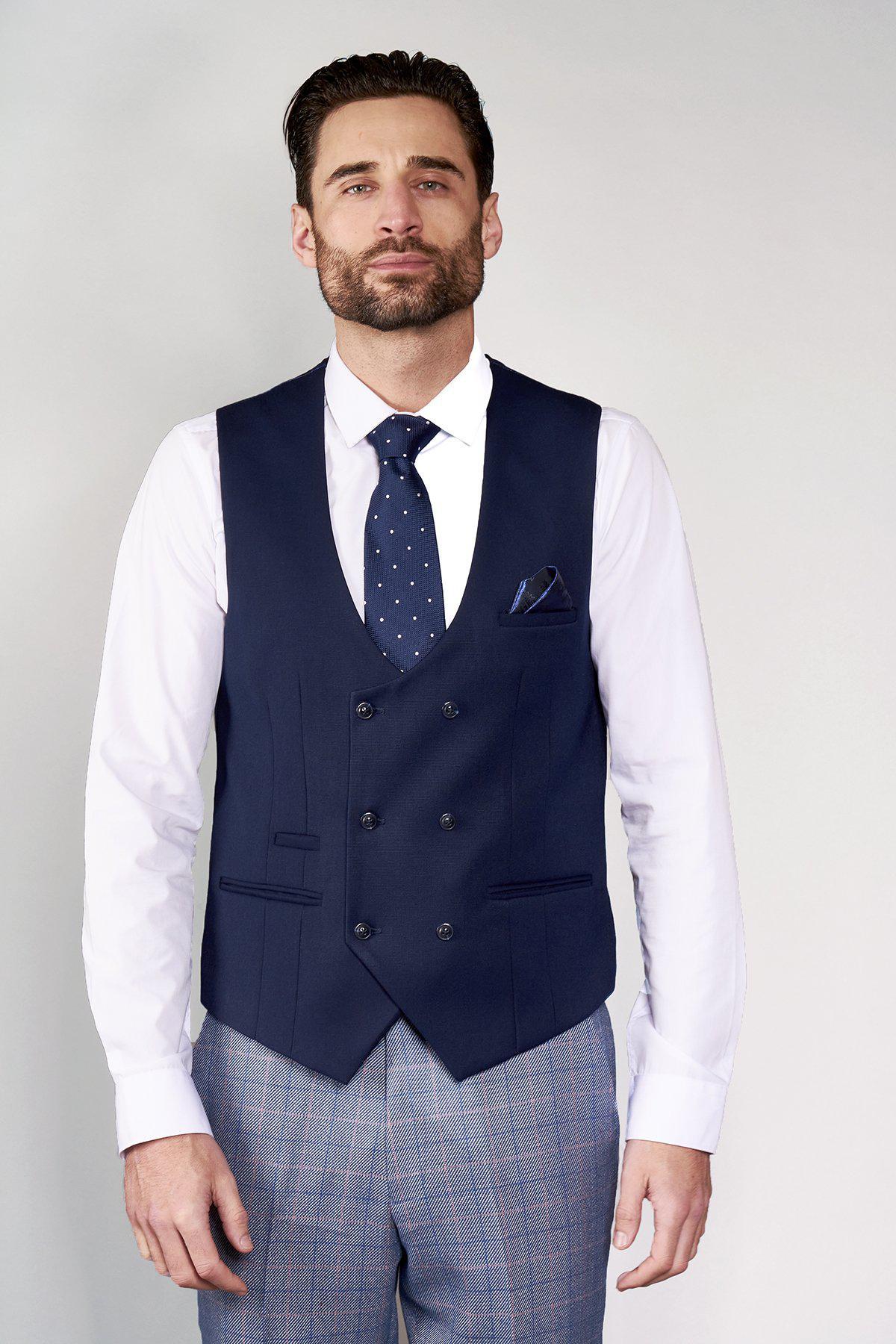 A man faces forward wearing the marcdarcy KELLY Blue Double Breasted Waistcoat, a white dress shirt, blue polka dot tie, and gray plaid trousers. He has slicked-back hair, light facial hair, and stands against a plain light background.