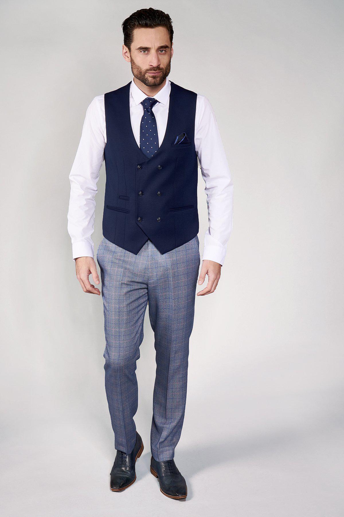 A man models the marcdarcy KELLY Blue Double Breasted Waistcoat with a white shirt, blue patterned tie, gray plaid trousers, and dress shoes against a light backdrop.