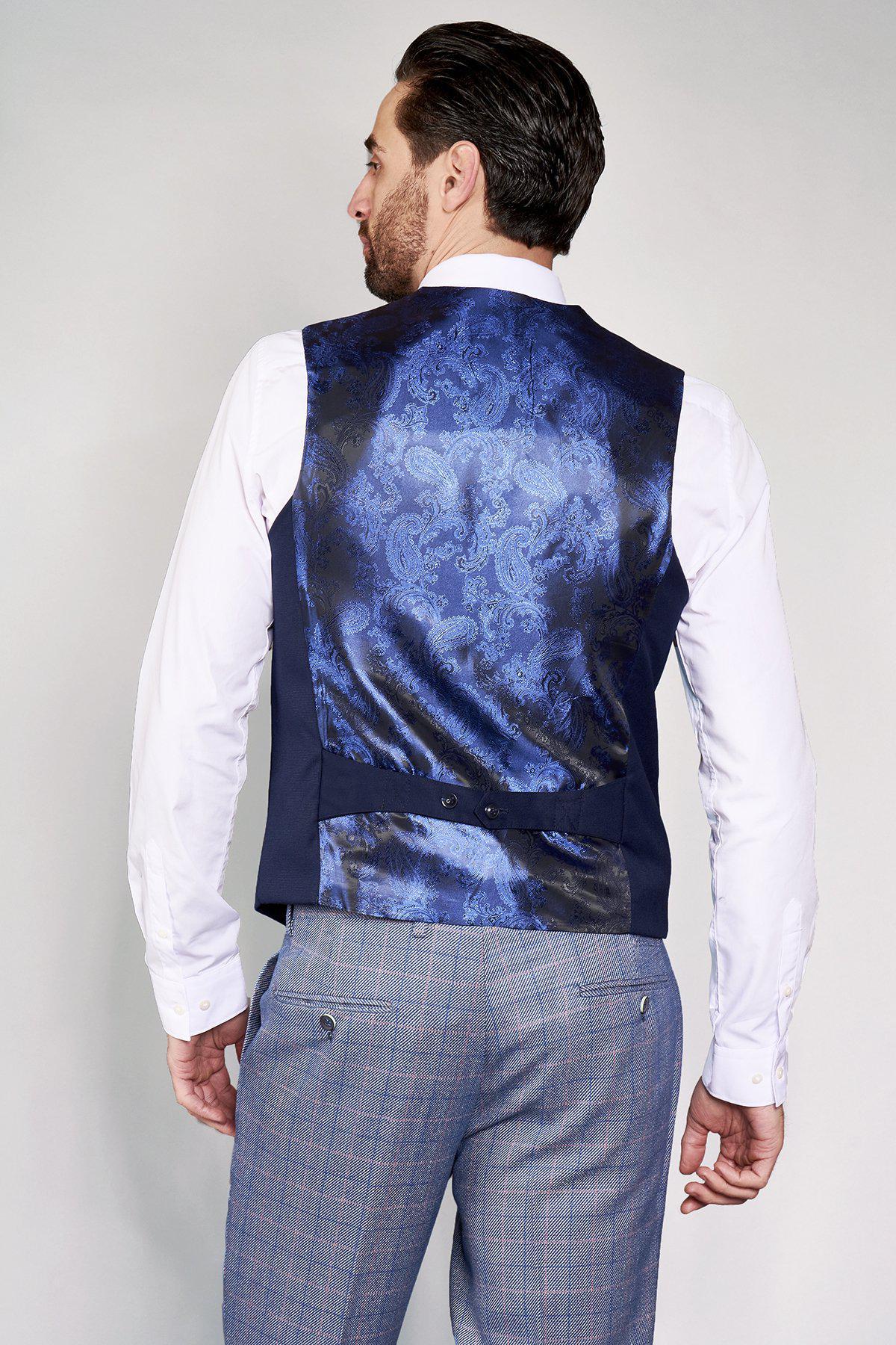 A man wearing a white dress shirt, marcdarcy KELLY blue double-breasted waistcoat, and light gray plaid dress pants stands with his back to the camera.