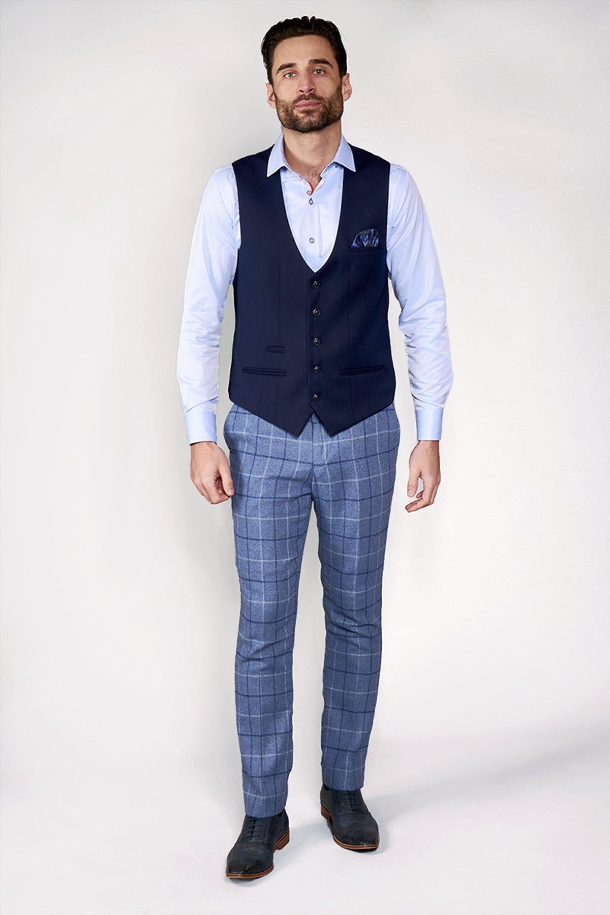 A man wears the marcdarcy KELLY Blue Single Breasted Waistcoat with a pocket square, light blue dress shirt, blue windowpane check trousers, and dark oxford shoes, standing against a white background.