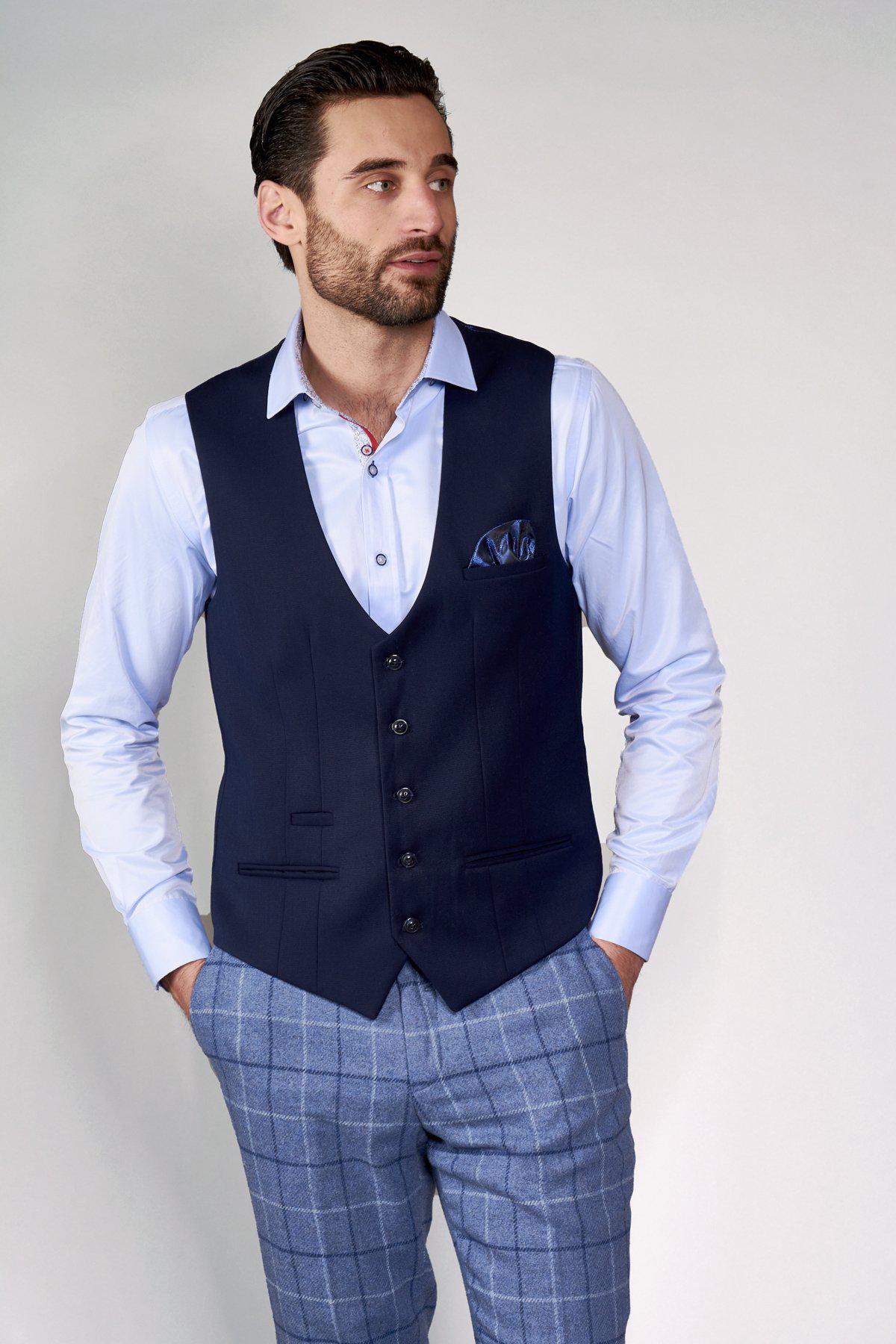 A man with dark hair and a beard wears a light blue shirt, blue plaid trousers, and the marcdarcy KELLY Blue Single Breasted Waistcoat. He stands with his hands in his pockets, looking to the side against a plain background.