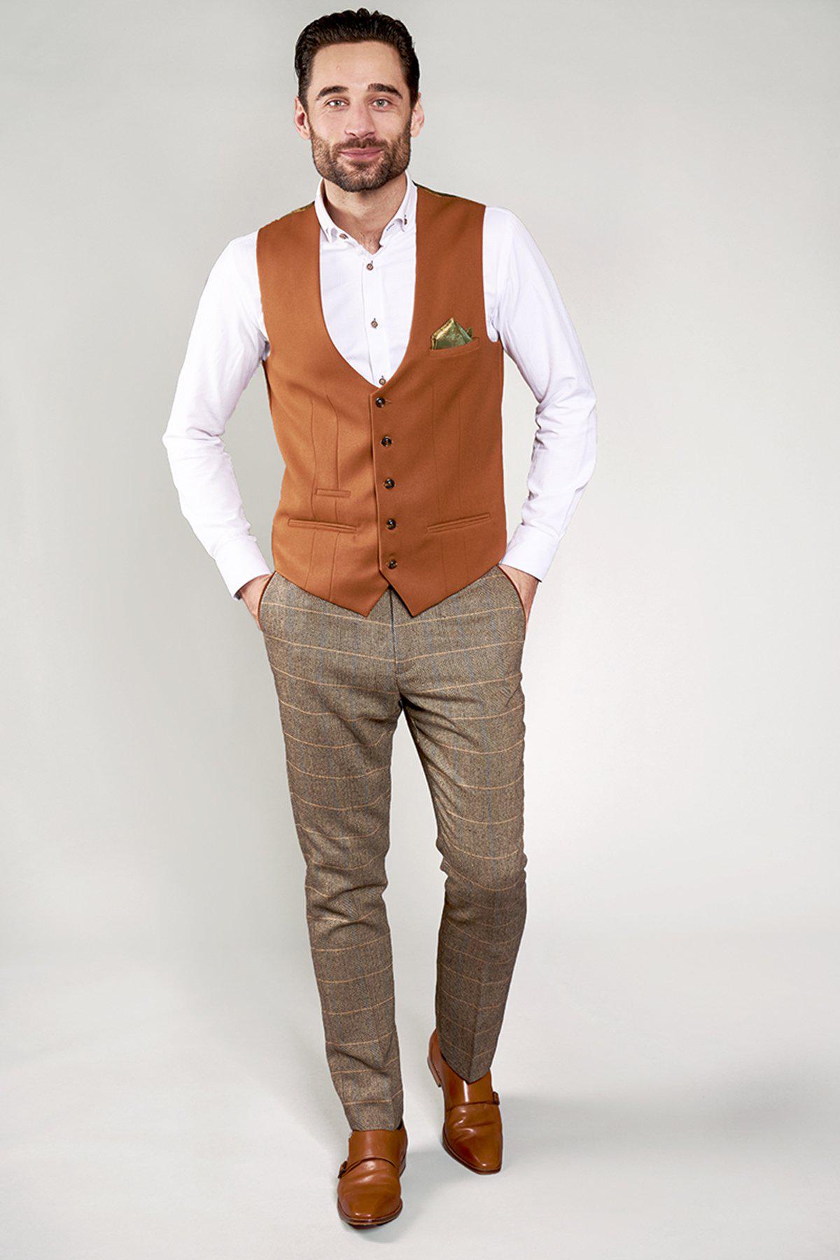 A man smiles at the camera, hands in pockets, wearing a white shirt, checkered brown pants, brown shoes, and the marcdarcy KELLY - Dark Tan Single Breasted Waistcoat with gold paisley lining against a plain light background.